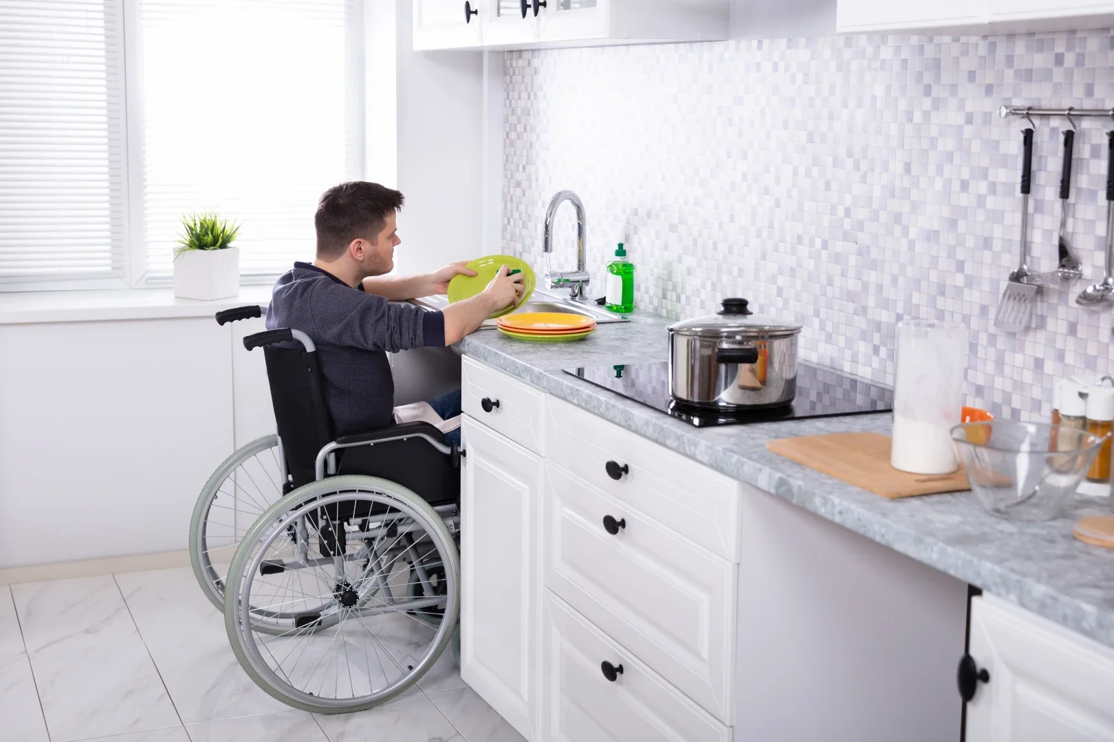 A man in a wheelchair washing dishes at a kitchen sink, with a stove, cutting board, and various kitchen items on the counter.