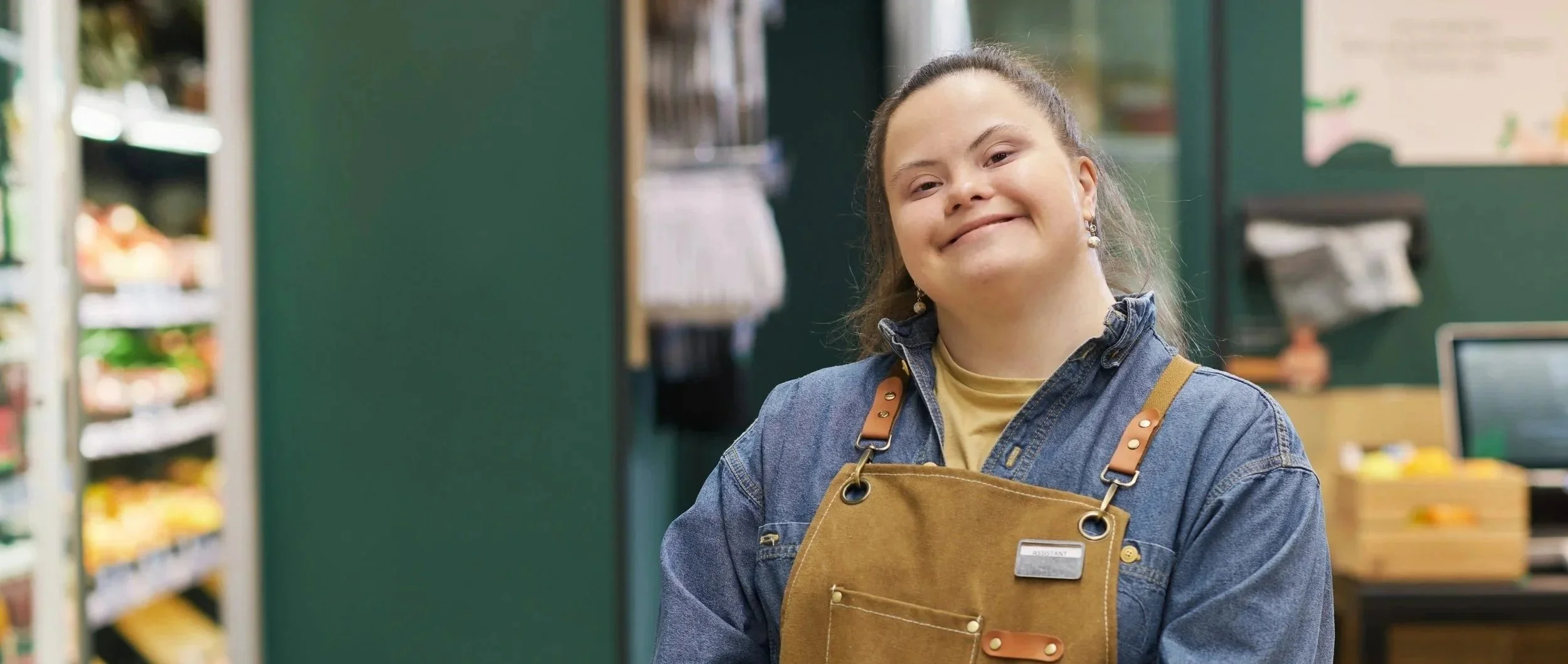 A smiling young woman in a grocery store, wearing a denim shirt and a tan apron with a name tag that says 'Assistant'.