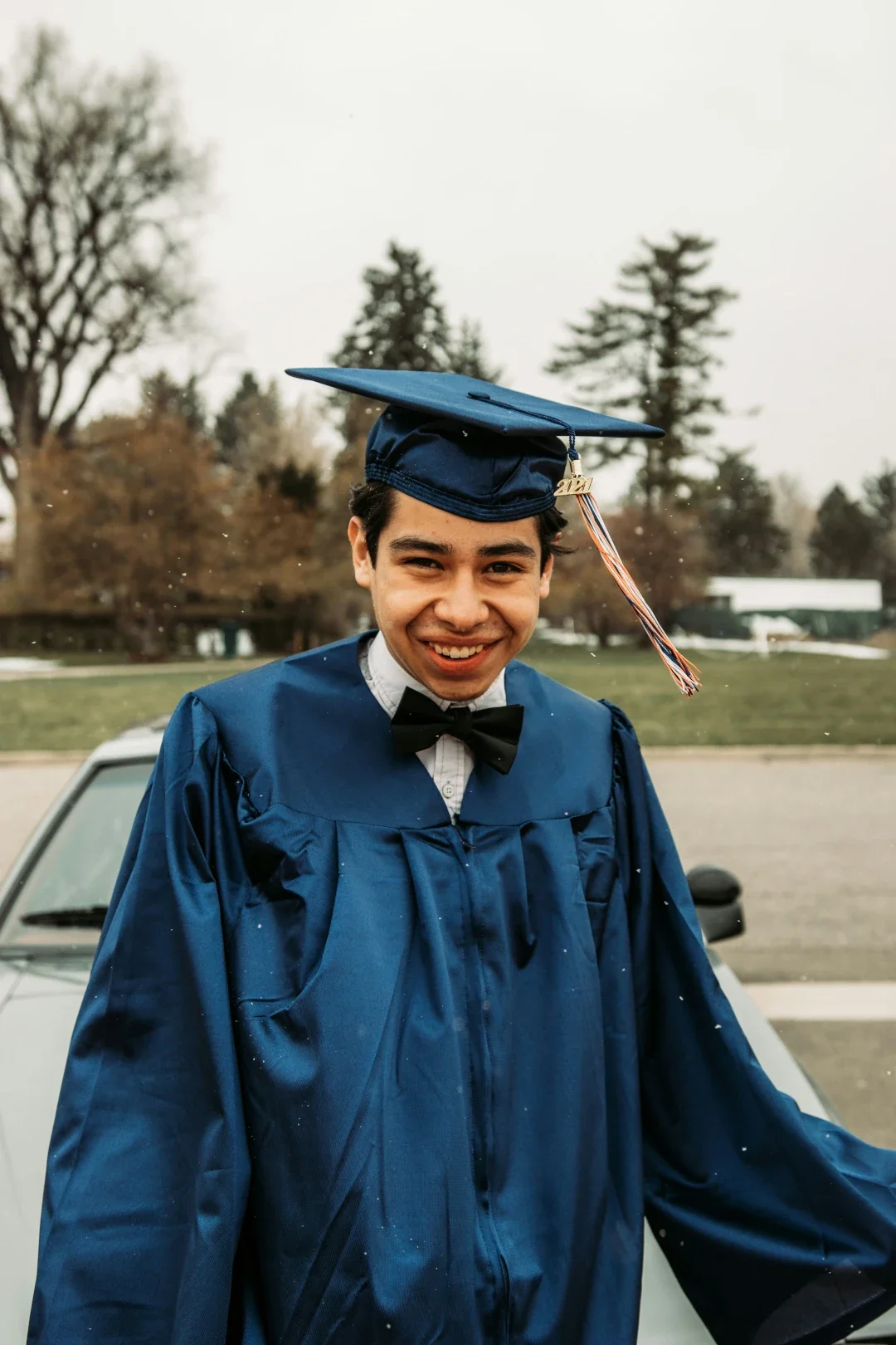 Young man wearing a blue graduation gown and cap with a tassel, smiling outdoors with trees and a car in the background.