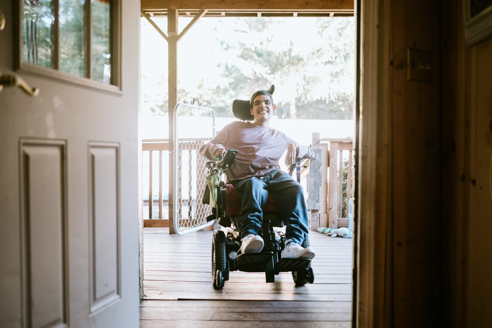 A man in a motorized wheelchair smiling and entering a house through an open door, with a wooden porch and outdoor scenery in the background.
