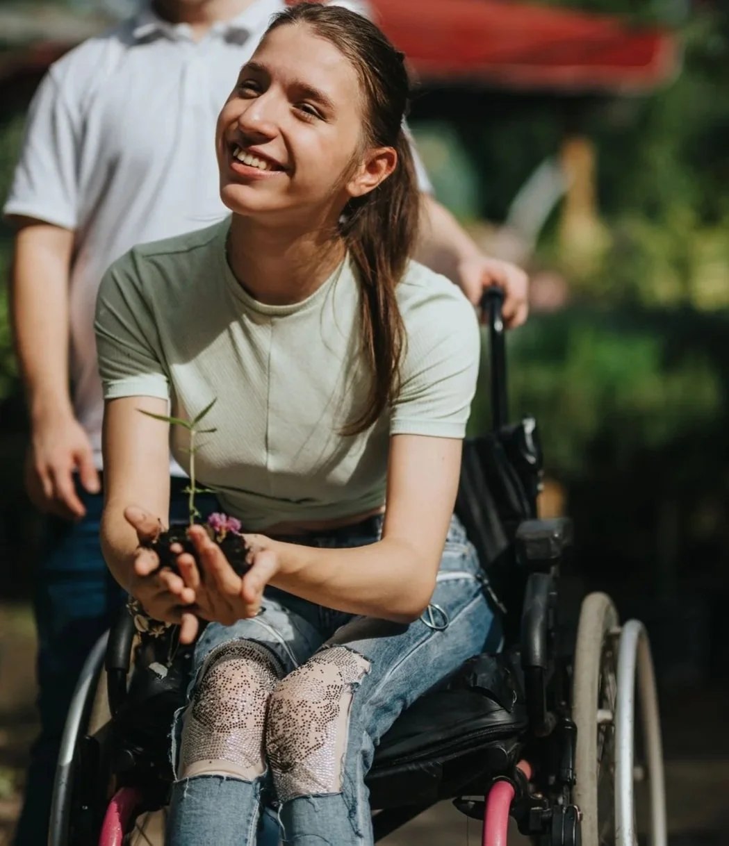 An occupational therapy client woman in a wheelchair smiling and holding a small potted plant, outdoors near a body of water with others in the background.