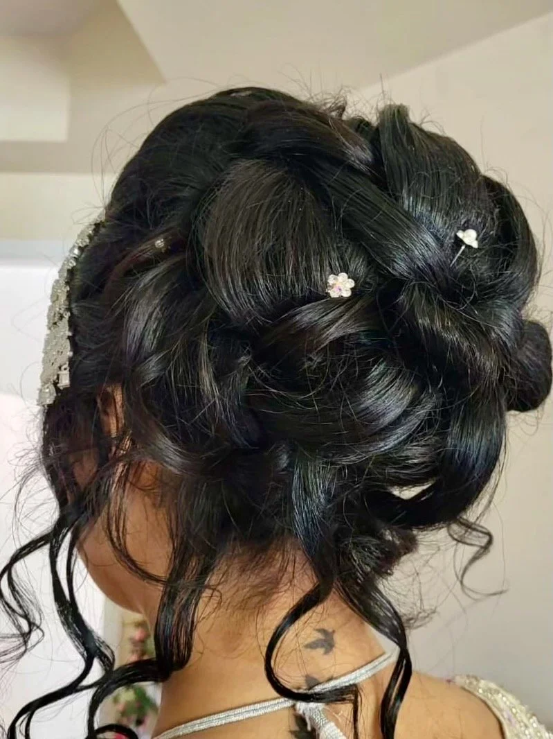 Close-up of an intricate updo hairstyle with curled sections of black hair, decorated with small white flower hairpins and a pearl headband, on a woman with a neck tattoo and wearing a silver strap dress.