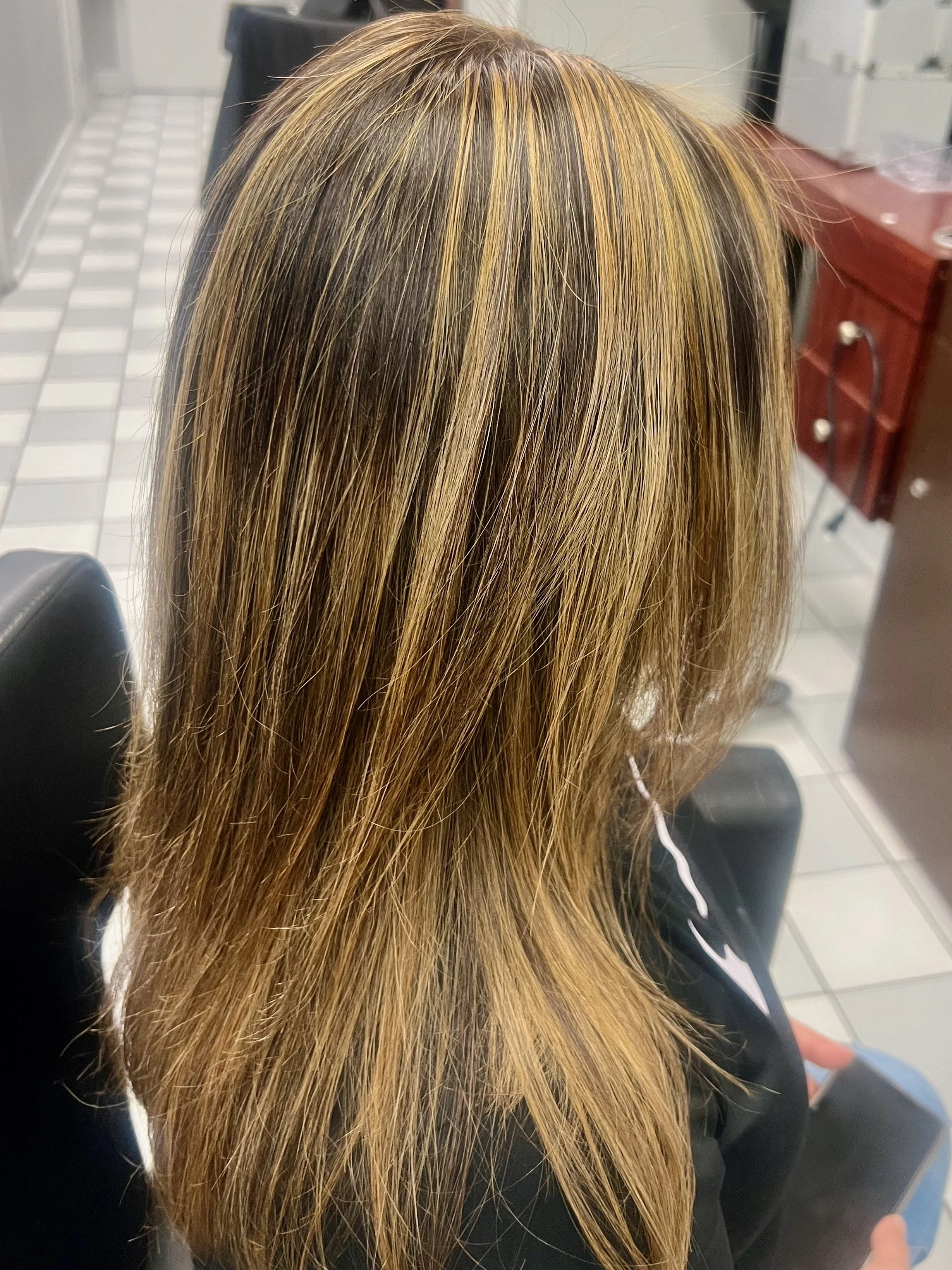 Overview of a person's head with medium-length, straight, light brown hair with blonde highlights, sitting in a salon or barbershop.