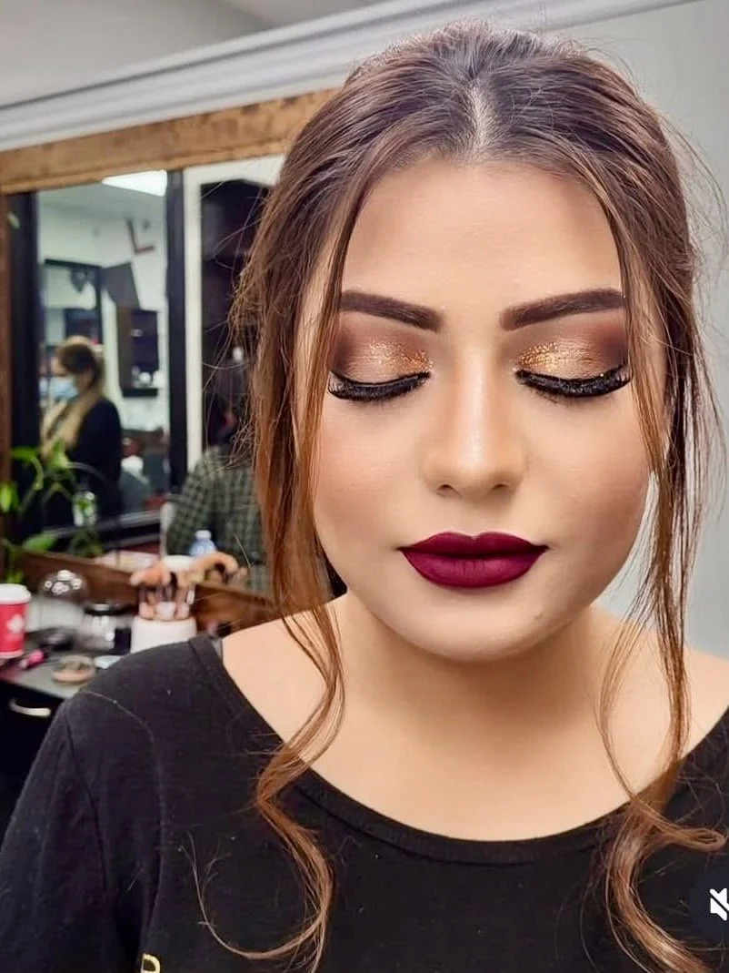 Young woman with makeup featuring gold eyeshadow and dark lipstick, posing with eyes closed in a salon or studio, with a mirror and another person visible in the background.