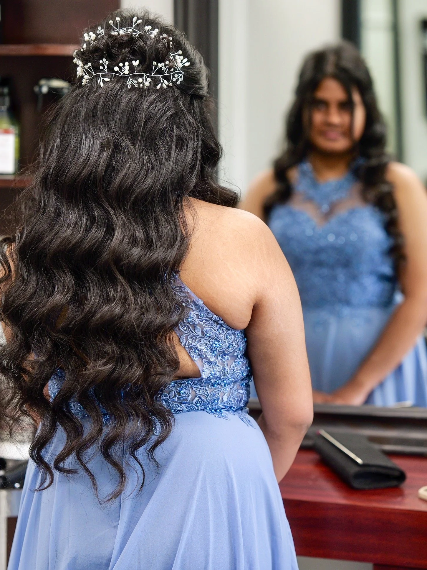 A woman in a blue dress with lace detail, long curly dark hair, wearing a white floral headpiece, looking at her reflection in a mirror.