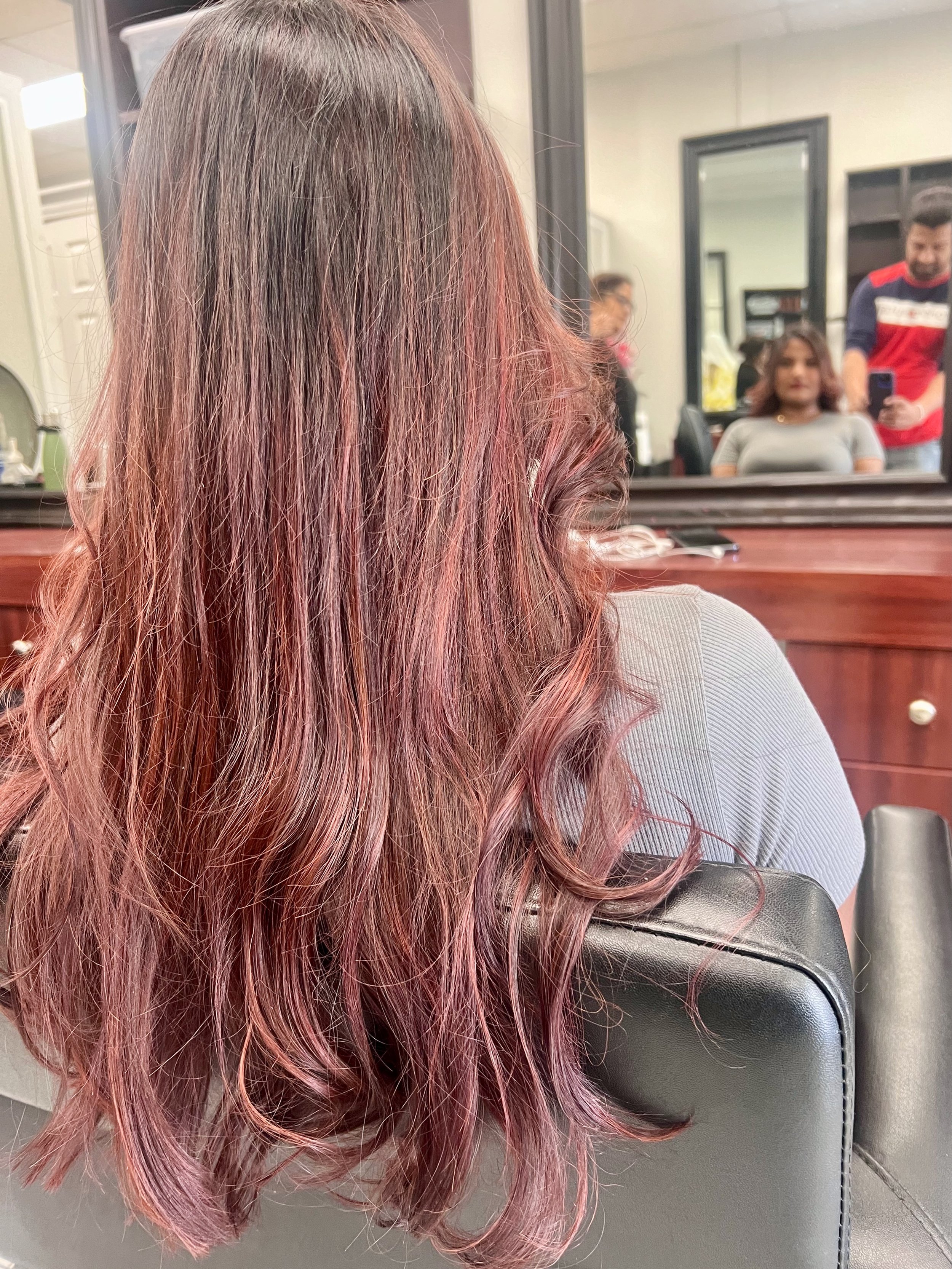 Person with long, wavy hair dyed in shades of pink and purple, sitting in a salon chair near a mirror, with two people visible in the background.
