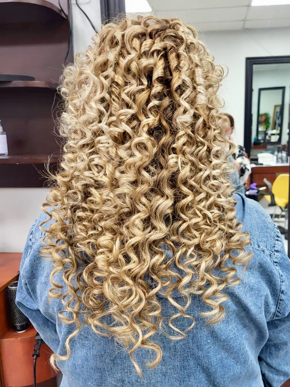 Back view of a woman with long, blonde, tightly curled hair, wearing a denim jacket, in a salon setting.