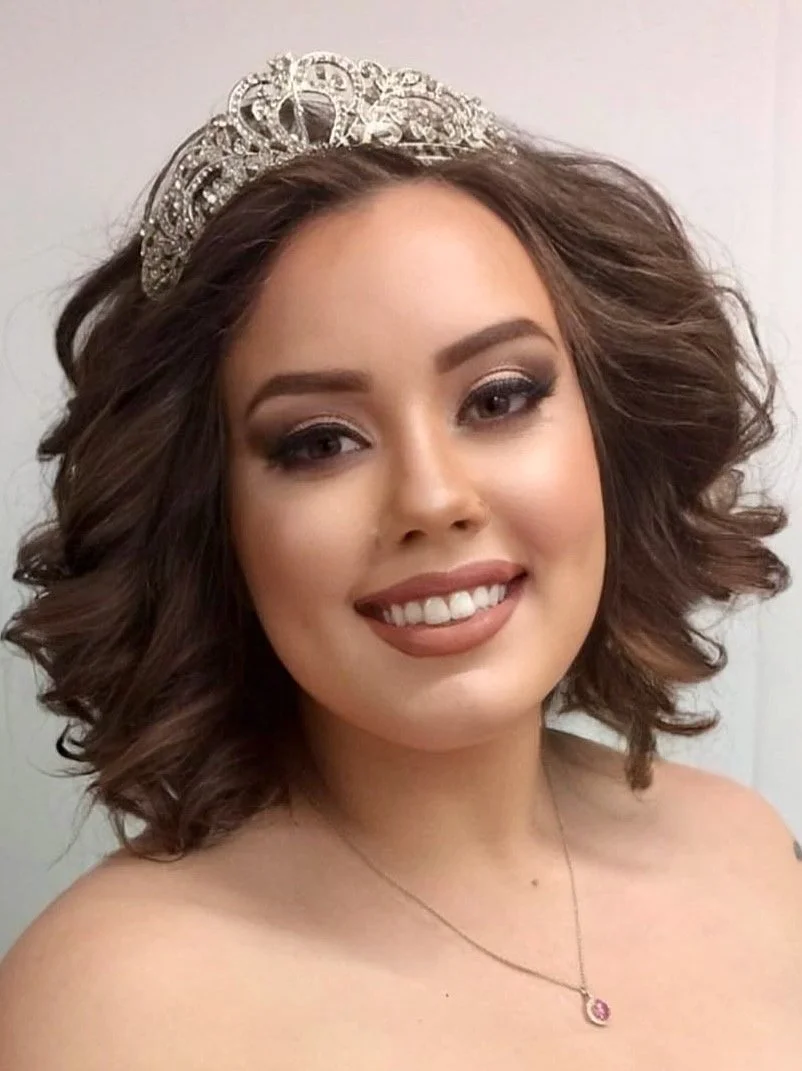 Young woman with wavy brown hair wearing a silver tiara and a pink gemstone necklace, smiling at the camera.