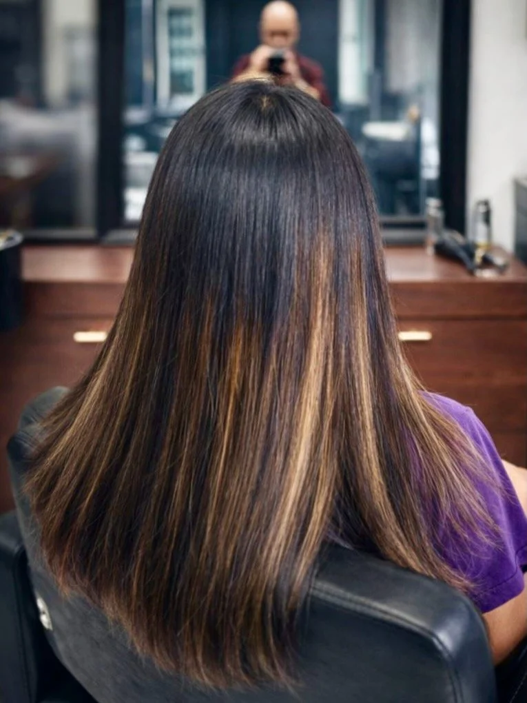 A woman with long, straight, dark brown hair with light brown highlights, sitting in a salon chair. A person in the background is taking a photo of her hairstyle in a mirror.