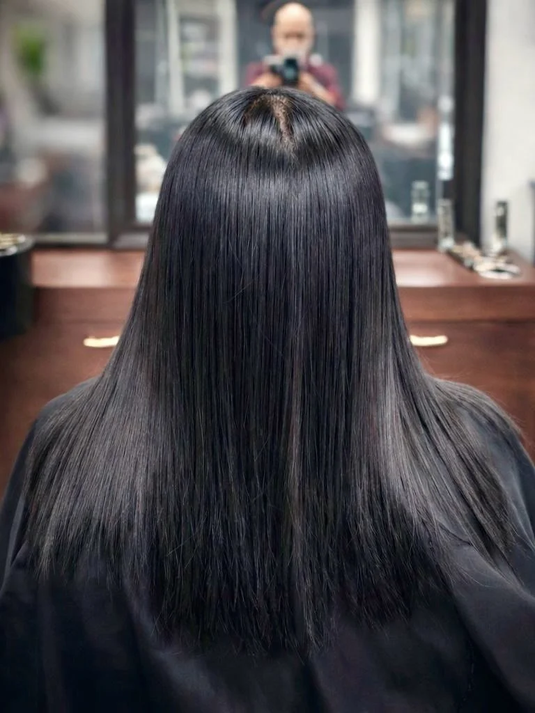 Person with straight, shiny, dark hair sitting in a salon chair, as a mirror reflects the person taking a photo.