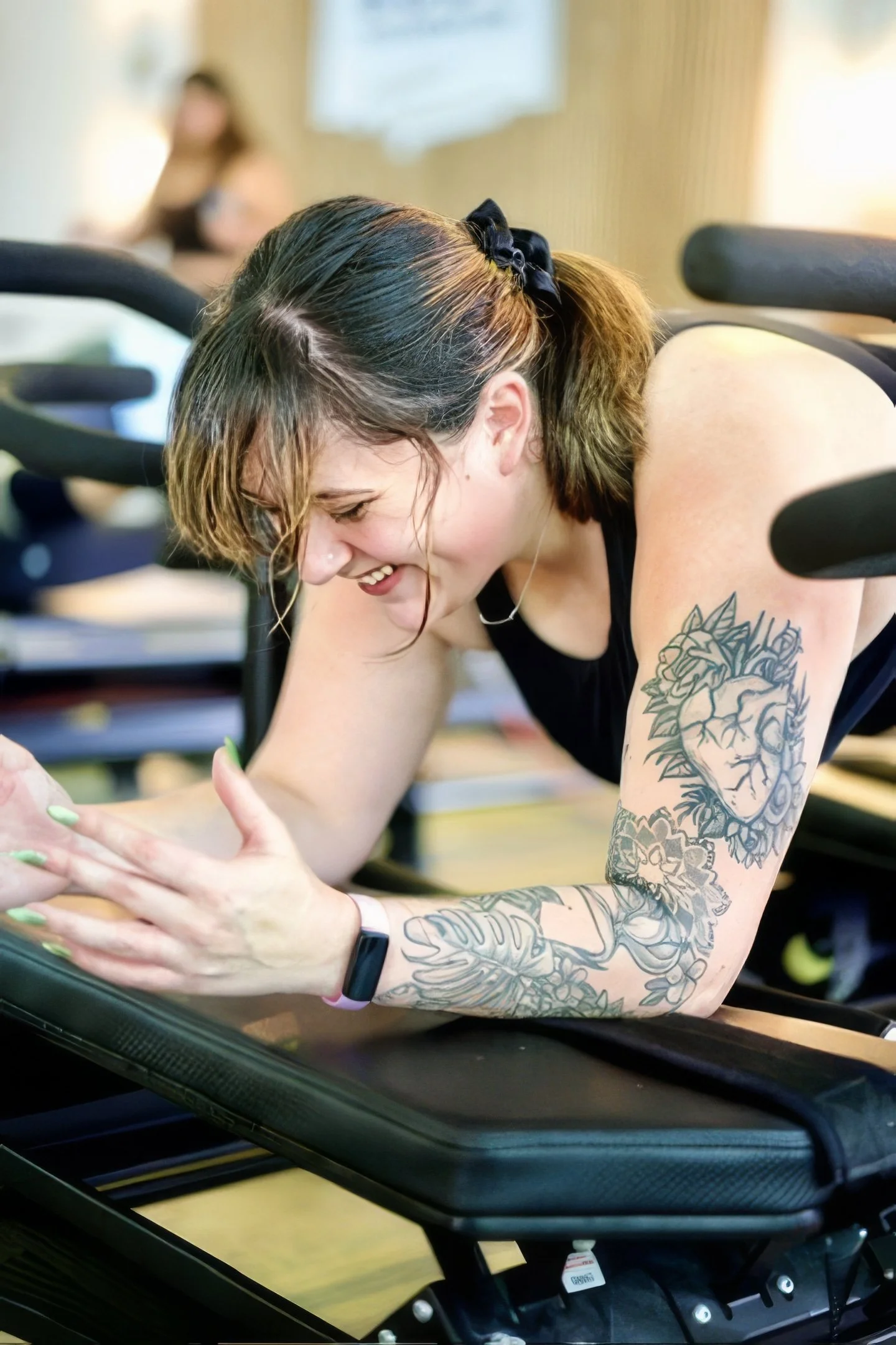 A woman with long dark hair and a black shirt sitting cross-legged on a piece of exercise equipment in a fitness studio.