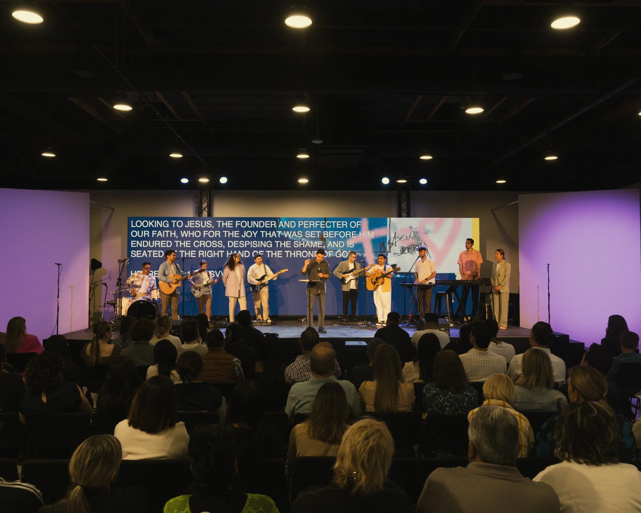 A church service or concert with a band performing on stage, including musicians playing guitars and keyboards, and a large screen displaying religious lyrics behind them. The audience is seated and watching the performance.