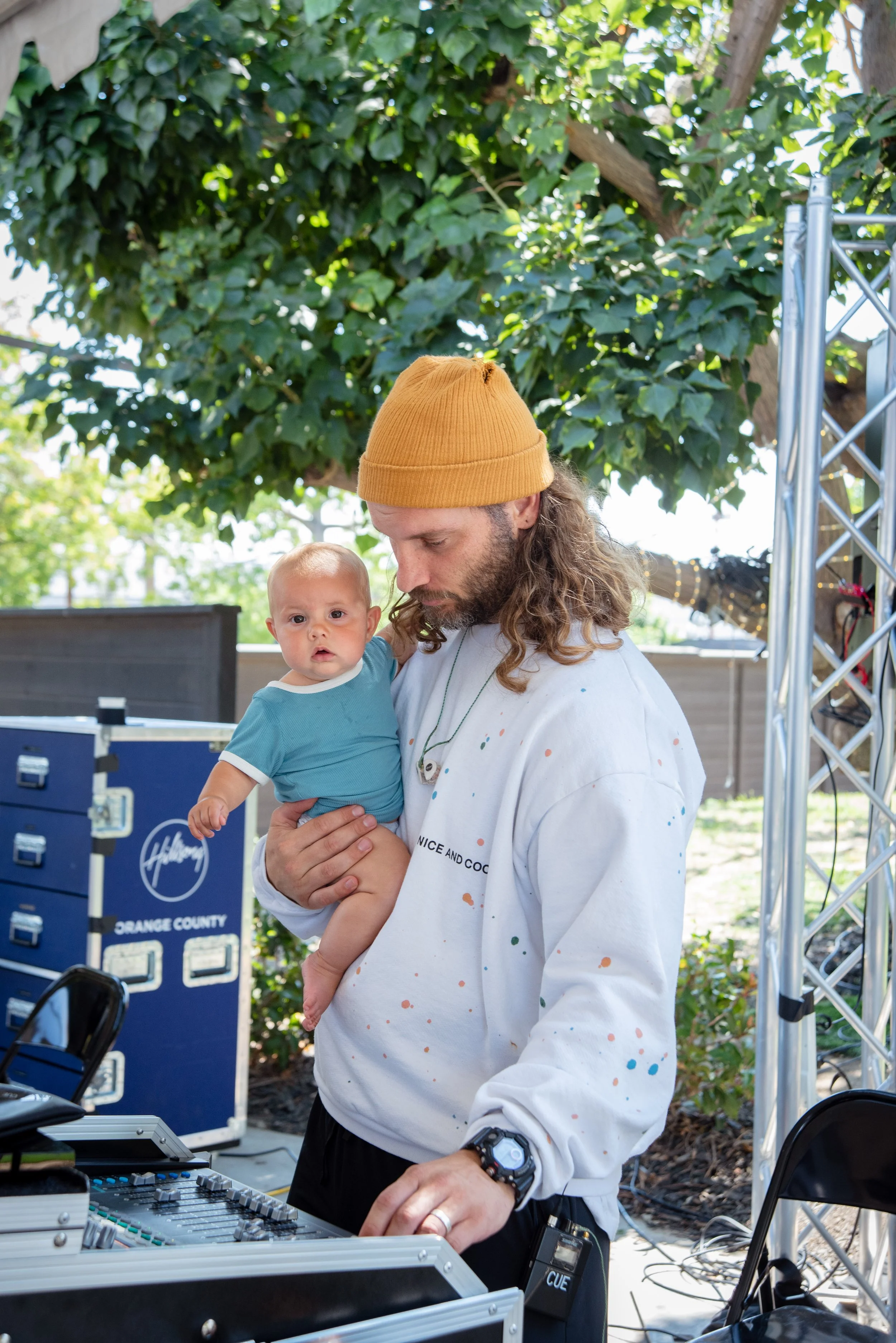 A man with long hair, wearing a yellow beanie and a white sweatshirt, holding a young child with a curious expression. The man appears to be working with audio equipment outdoors, under a large green tree, with some equipment and a blue box labeled 'Orange County' nearby.