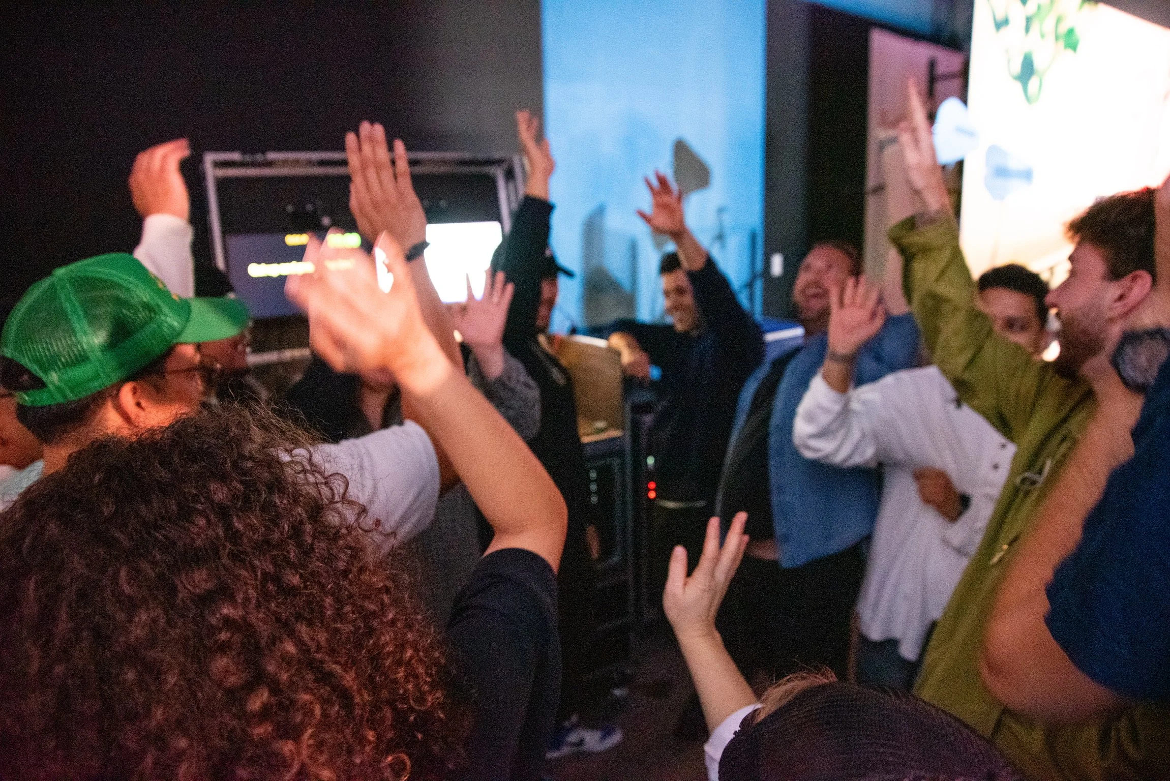 Group of people celebrating with raised hands in a dimly lit indoor setting, possibly at a party or concert.