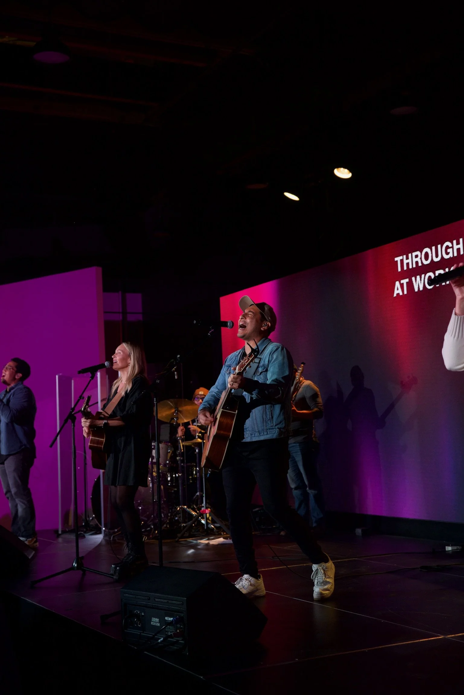 Band performing on stage with microphones and musical instruments, with a presentation screen in the background displaying text.