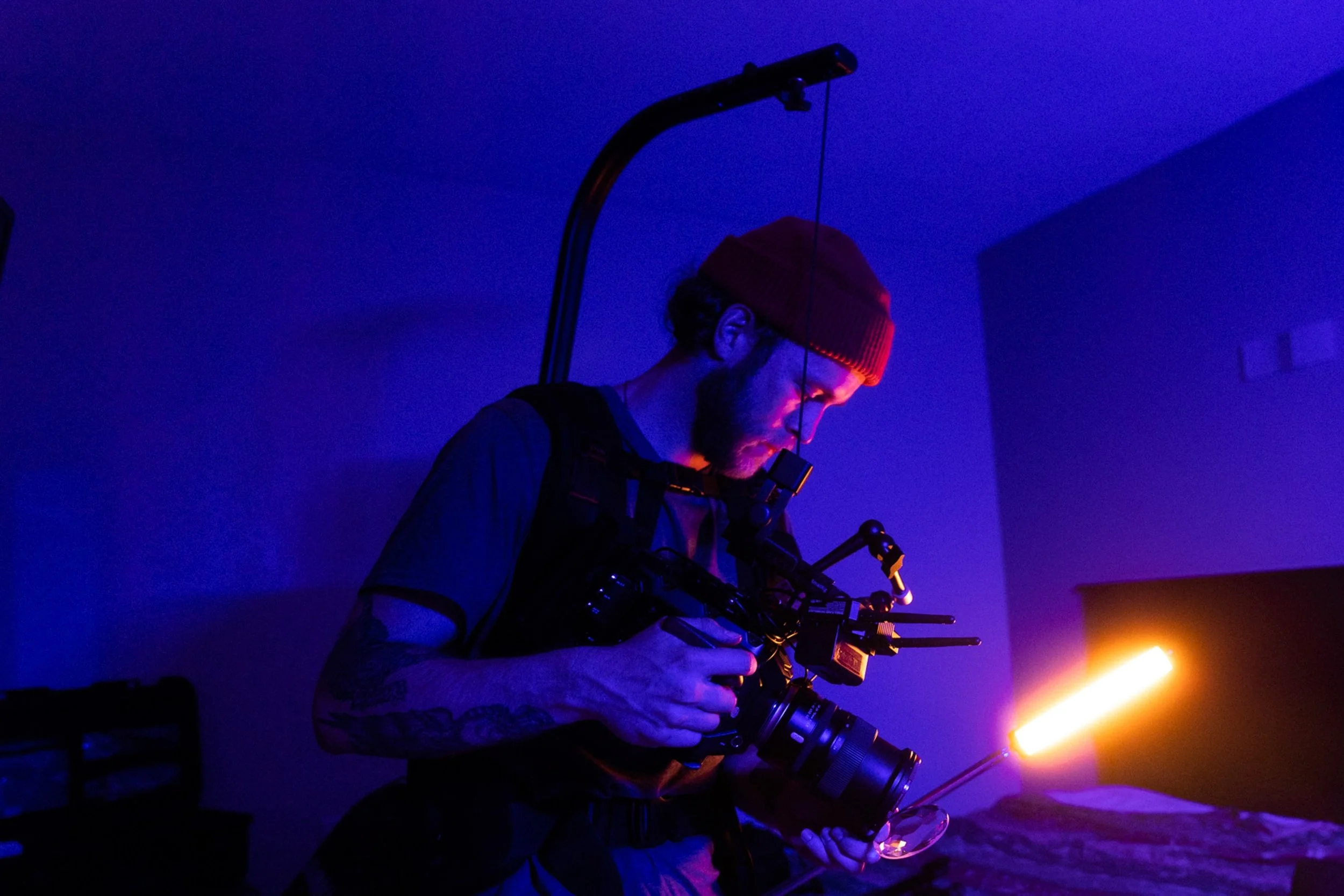 A man with a beard and tattoos wearing a red beanie and black clothing, using a camera rig with a mounted long light in a dark room with blue and purple lighting, looking at a small object in his hand.