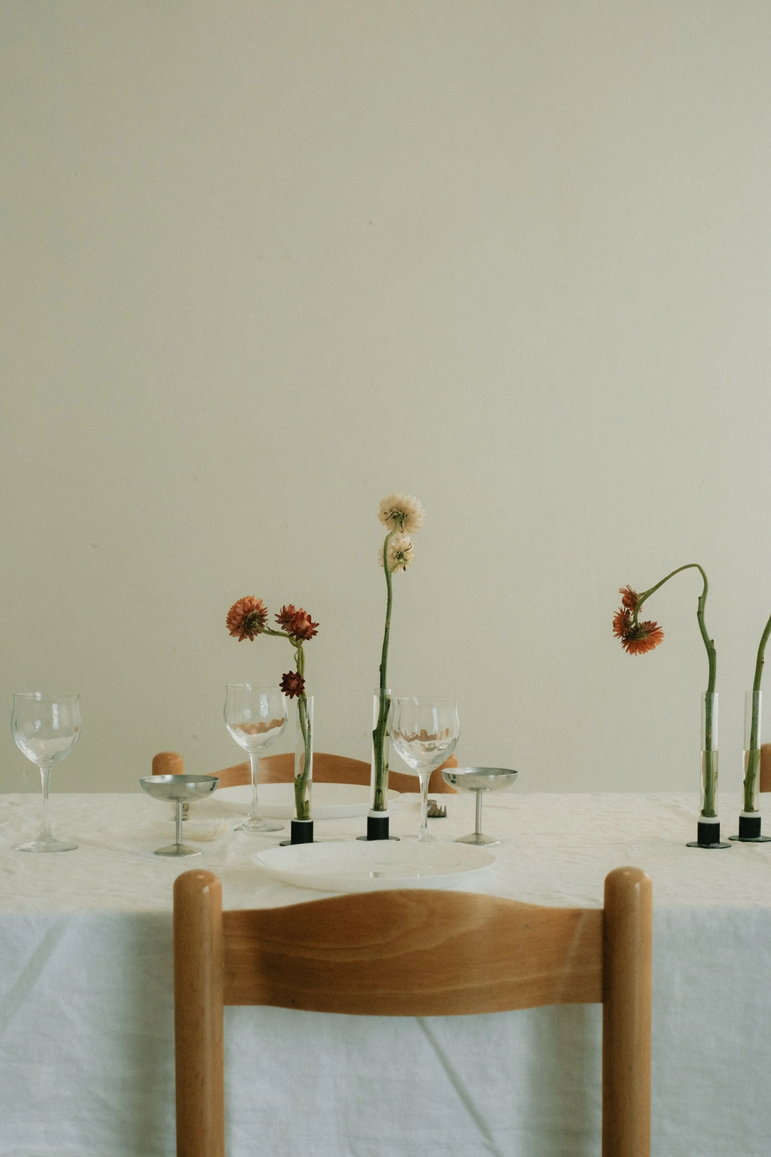 Table setup with four tall glass vases holding flowers, surrounded by wine glasses and small bowls, on a white tablecloth in a dining room.