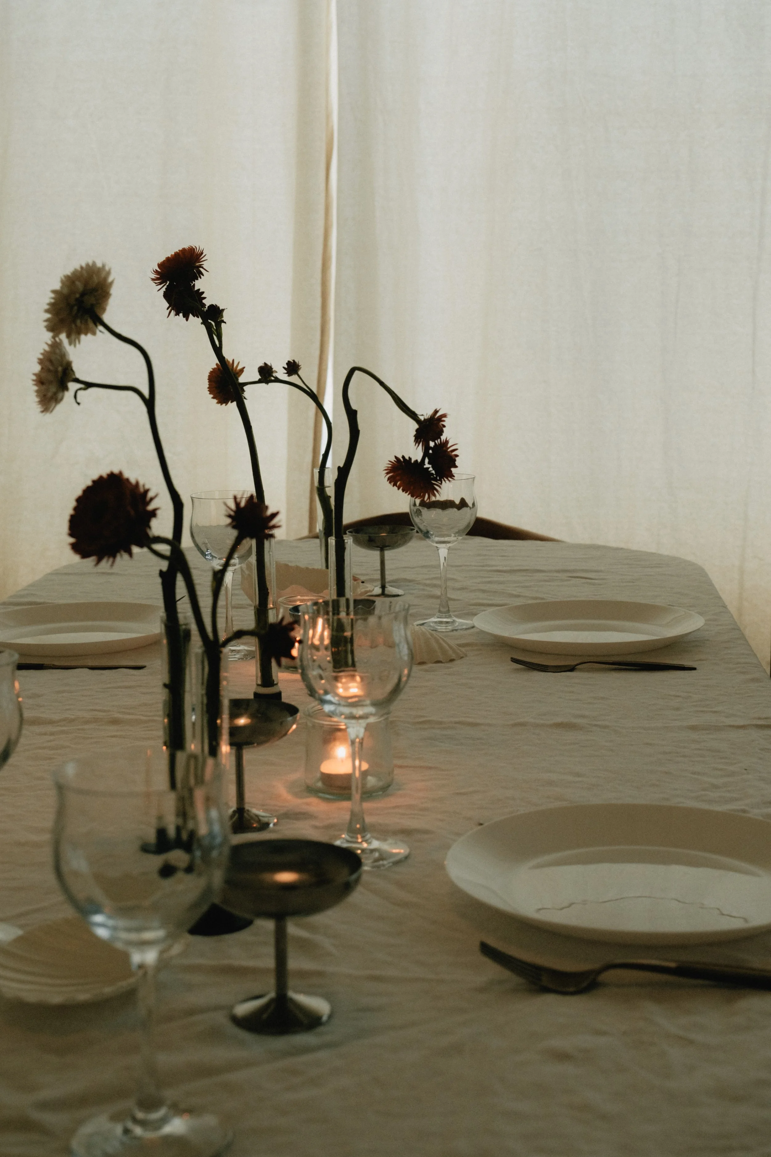 A dining table set with multiple wine glasses, some with red wine, and white plates. Vases with wilted flowers and candles add to the dimly lit, elegant table setting. A light curtain is in the background.