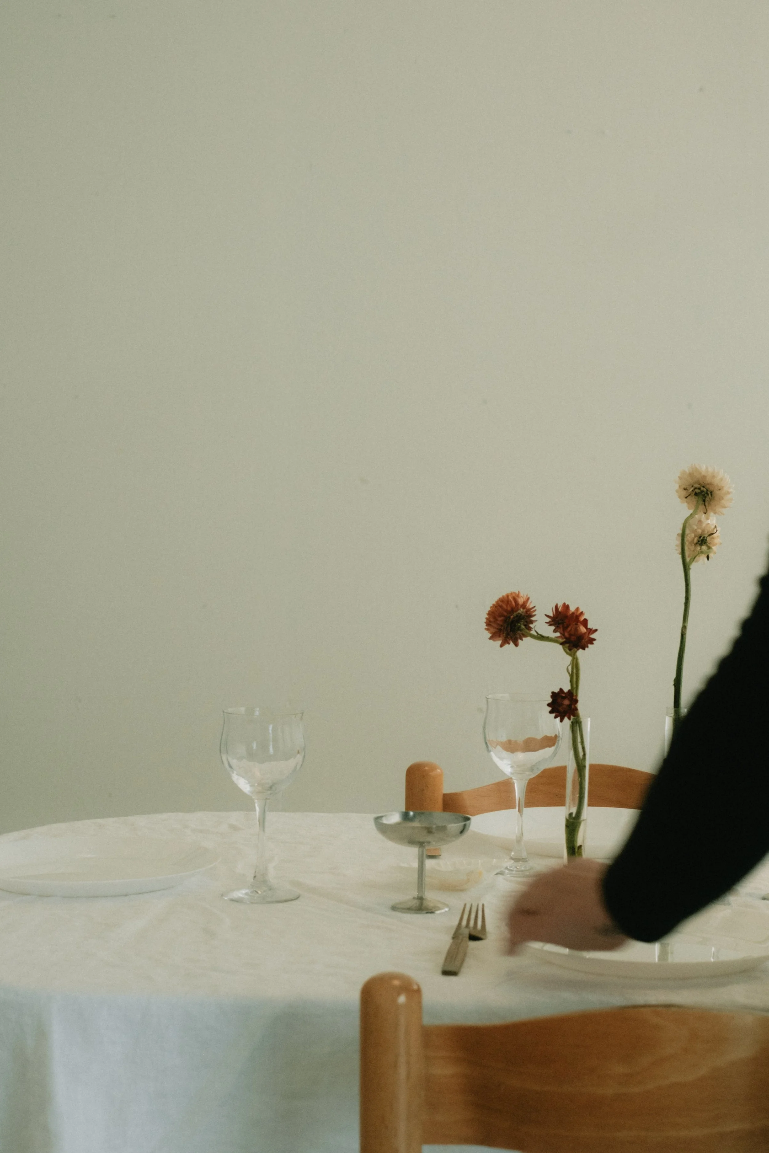 A table set with empty wine glasses, a plate, cutlery, and a vase with flowers, next to a wooden chair. A person's hand is reaching for something on the table.