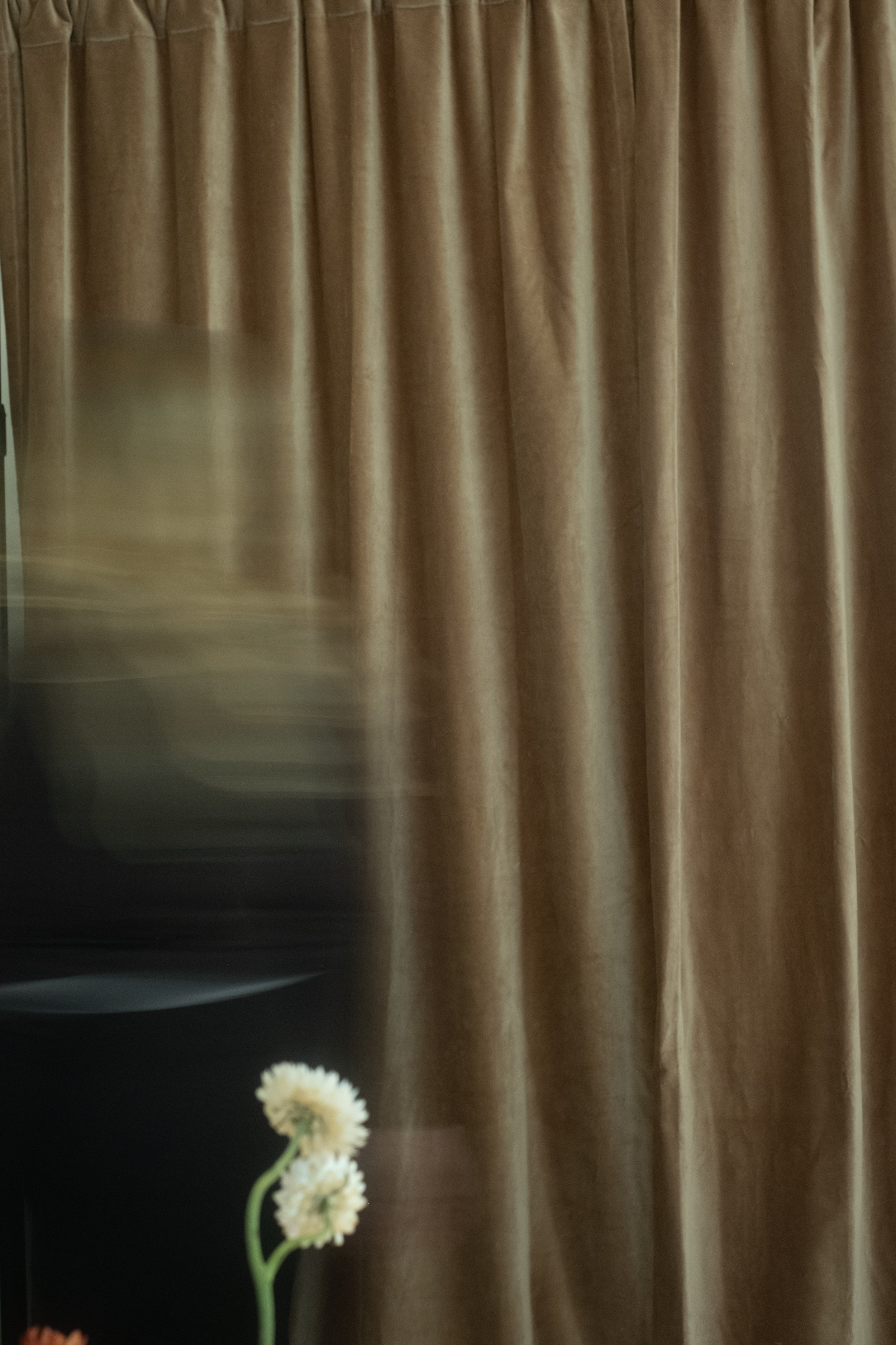 A brown curtain with vertical pleats. Part of a dark surface with a white flower in the lower left corner is visible in the foreground.
