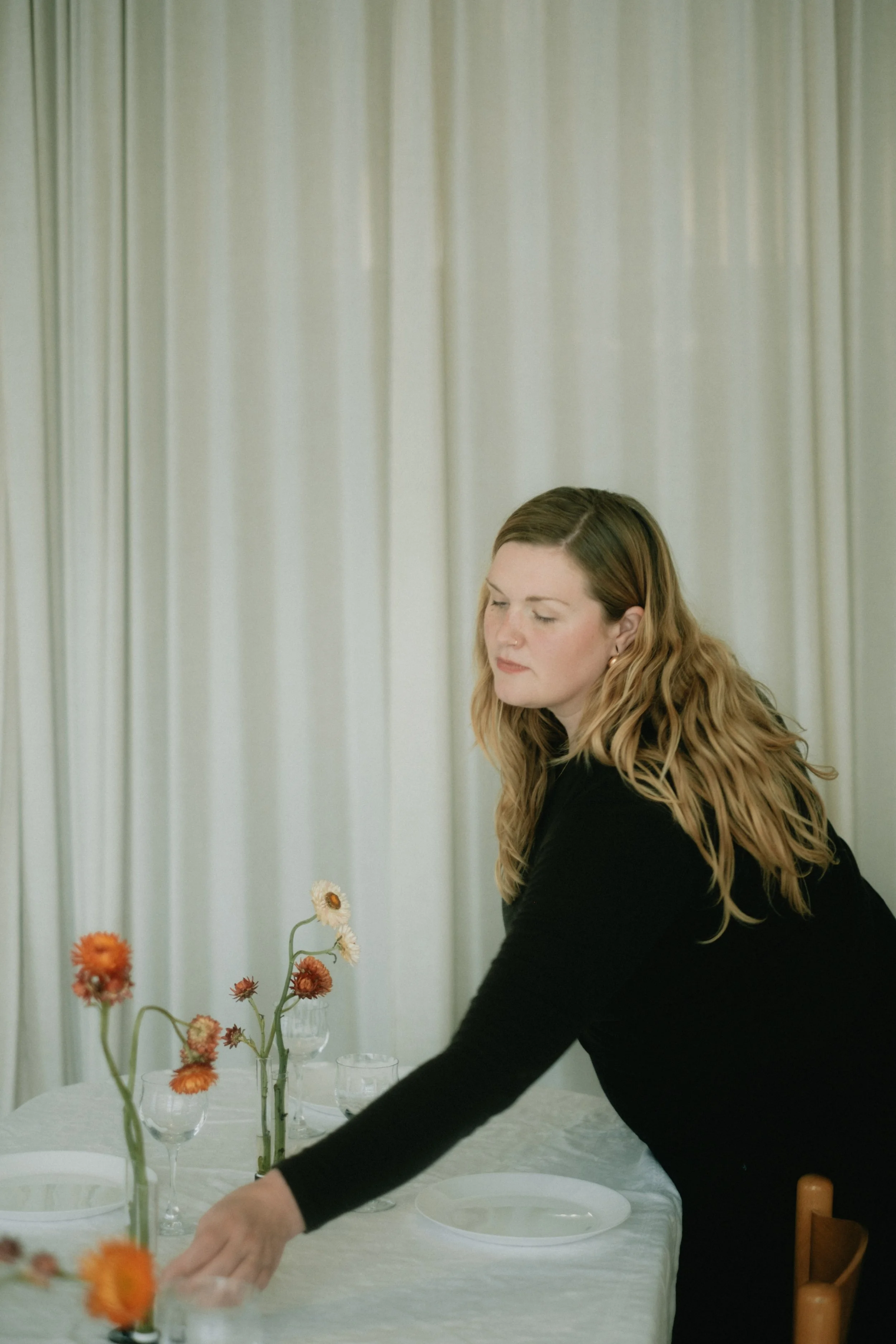 A woman with long, wavy brown hair arranging orange and white flowers on a table with a white tablecloth, in front of a gray curtain.