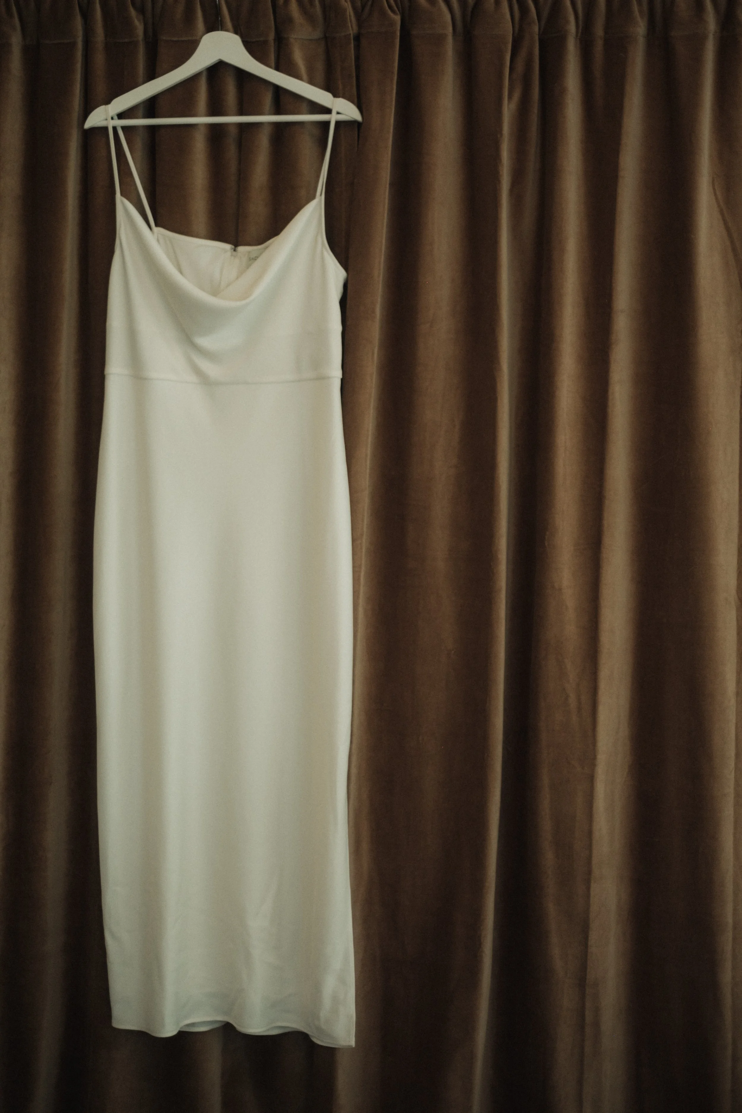White dress hanging on a white hanger in front of brown curtains.