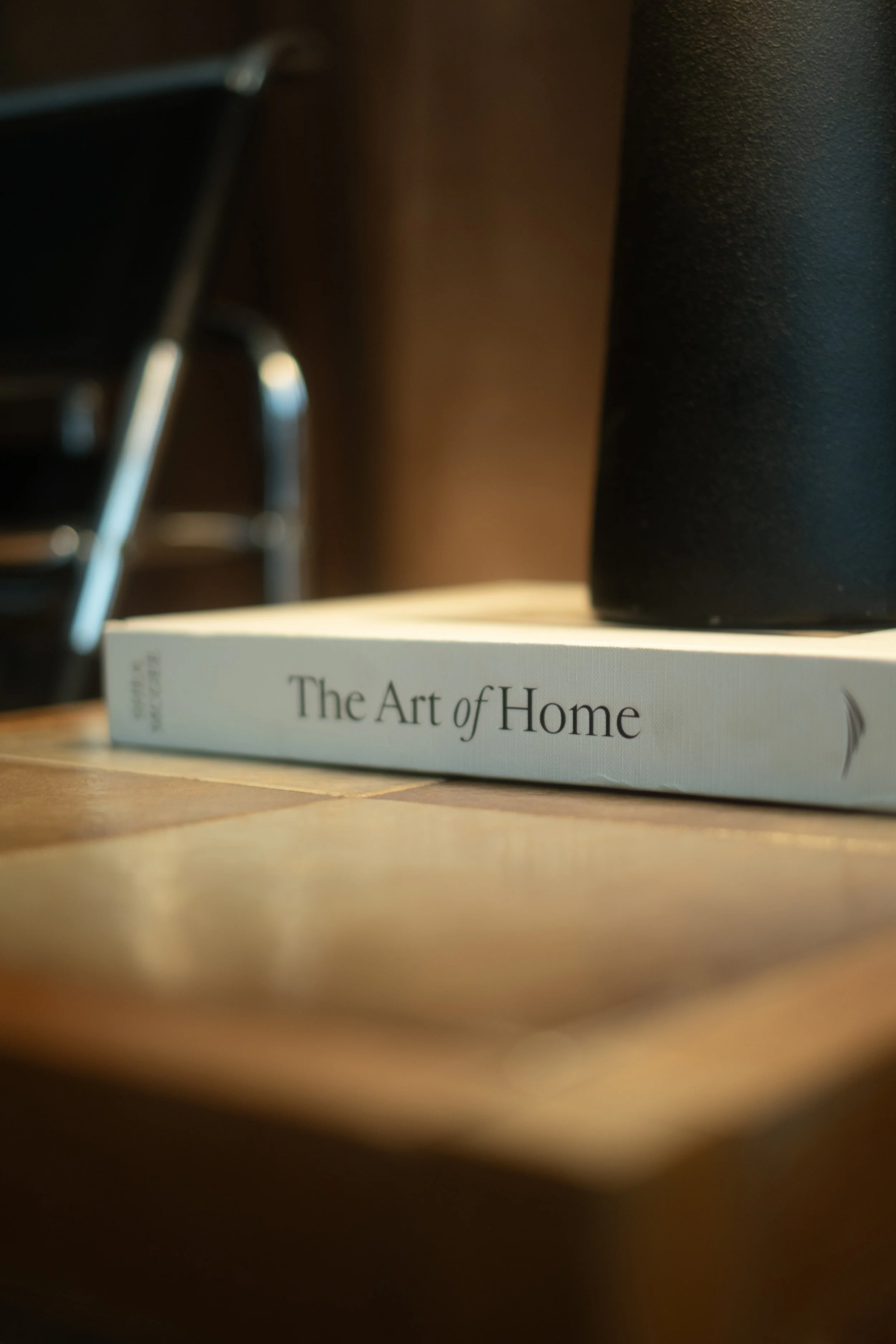 Close-up of a book titled 'The Art of Home' on a wooden surface, with blurred objects including a black container and possibly a piece of furniture in the background.
