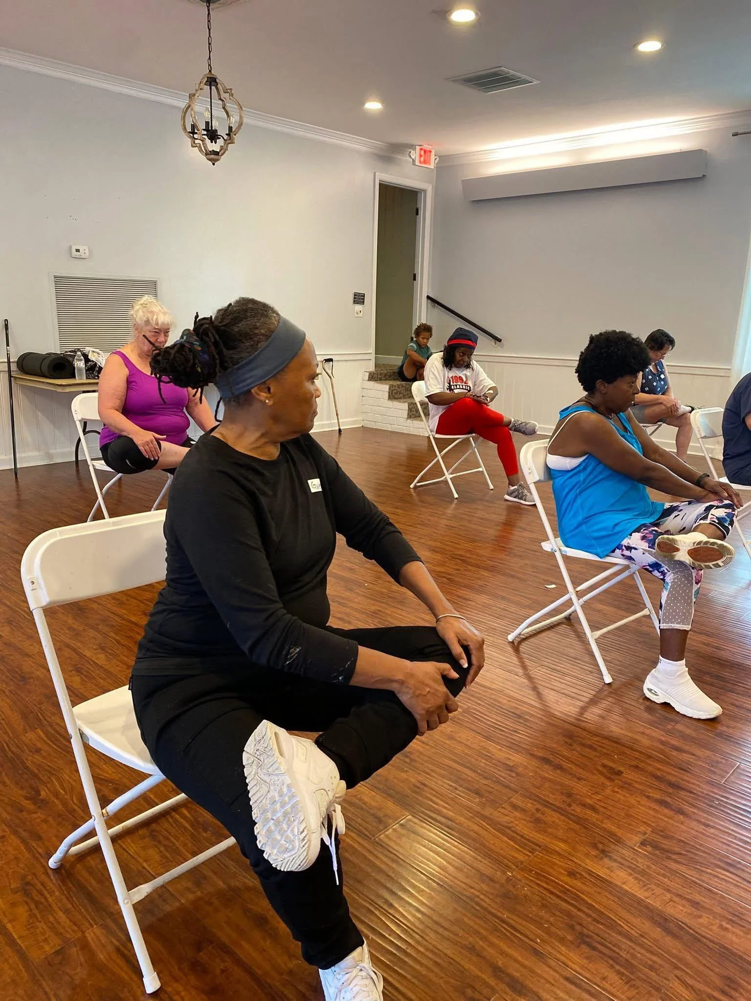 Woman performing a yoga pose indoors, kneeling with one arm bent and fist near her face, wearing a pink shirt and purple pants.