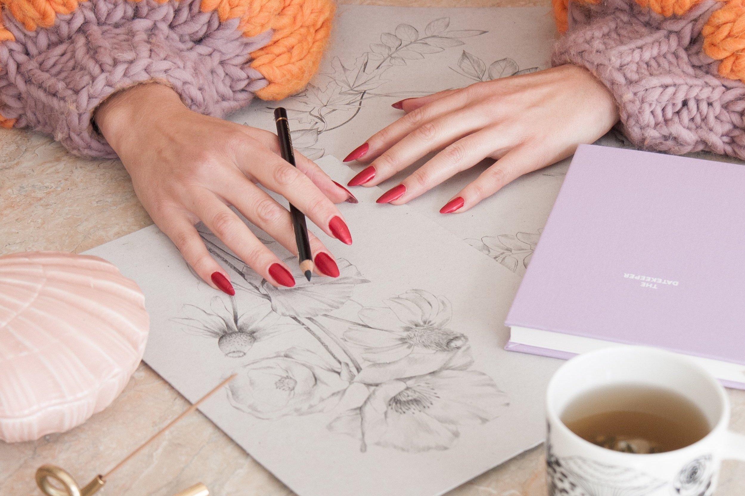 Person with red painted nails sketching floral designs on paper with a pencil, surrounded by a pink shell-shaped object, a purple notebook, and a cup of tea.