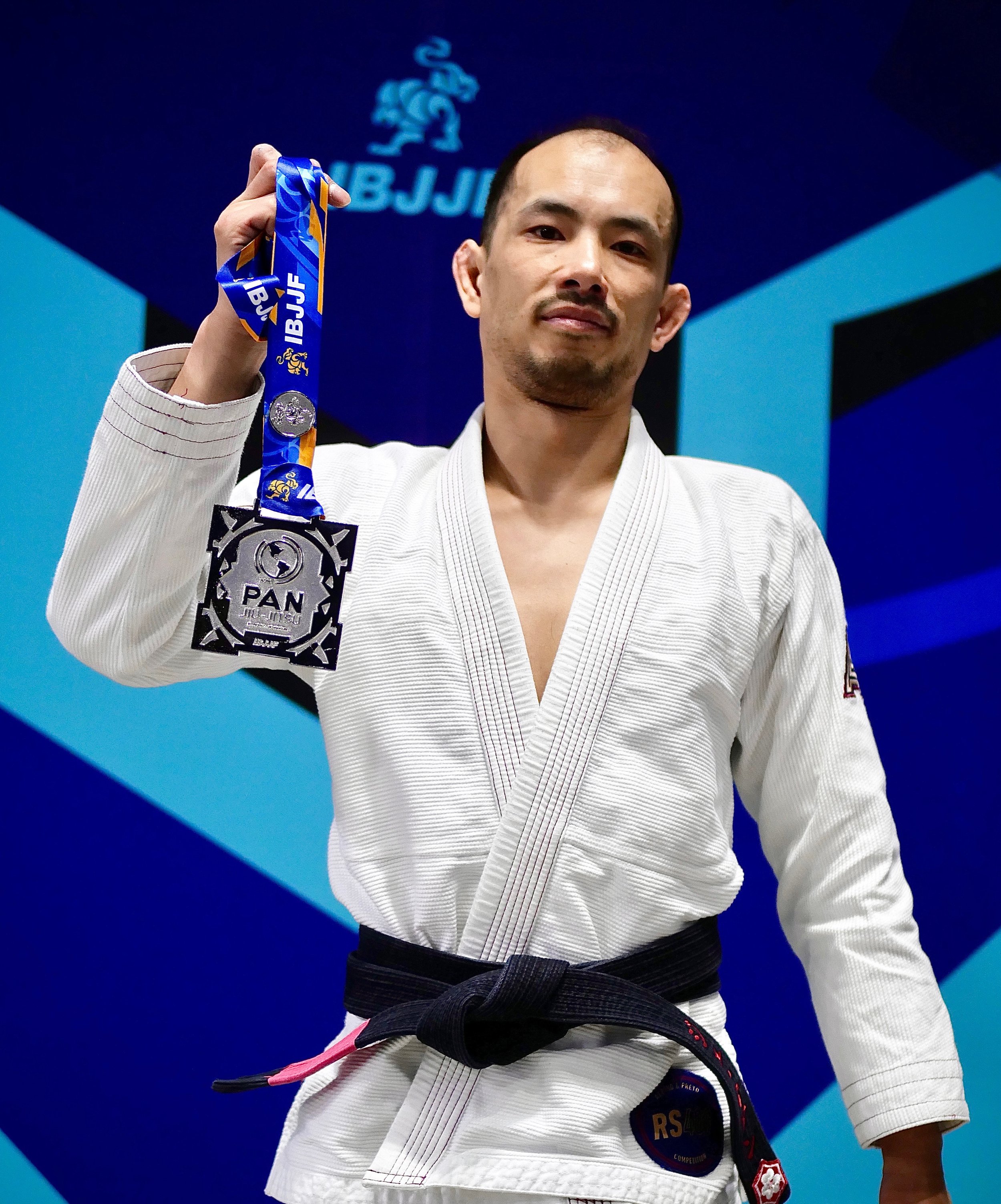 A martial artist in a white gi holding a silver medal with the IBJJF logo and PAN inscription, signifying a placing in a Brazilian Jiu-Jitsu competition.