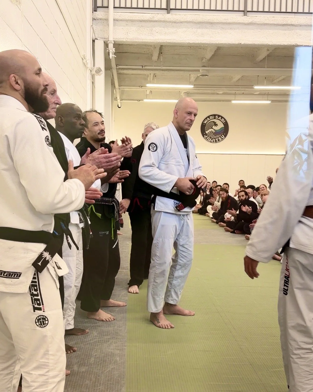 BJJ students and instructors gathered in a dojo listening to an instructor, who is in gi, holding his black belt, with seated students in the background and a Wabi-Sabi Jiu Jitsu sign on the wall.