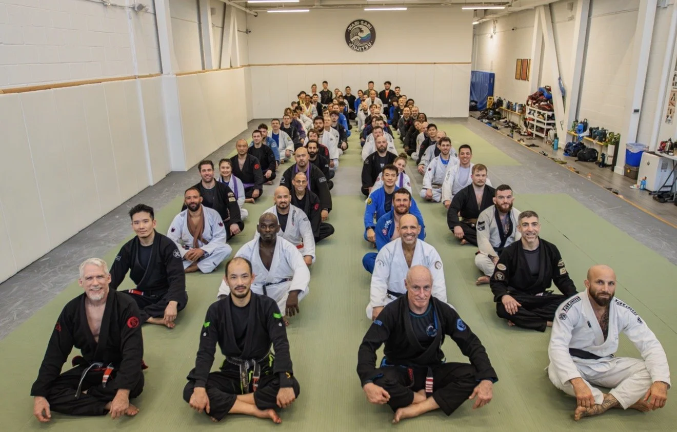 Group of martial artists in traditional uniforms practicing Brazilian Jiu-Jitsu on mats in a training facility.