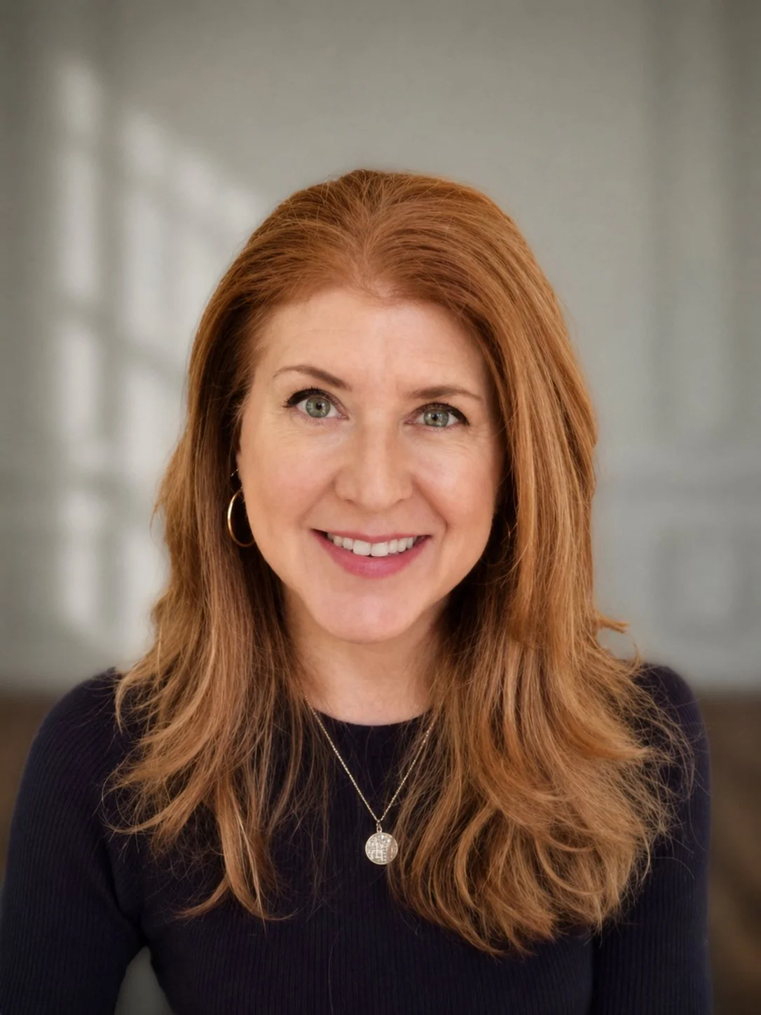 Close-up photo of Elise Arnoult Miller with reddish hair, green eyes, wearing a black top, gold hoop earrings, and a silver pendant necklace, smiling.