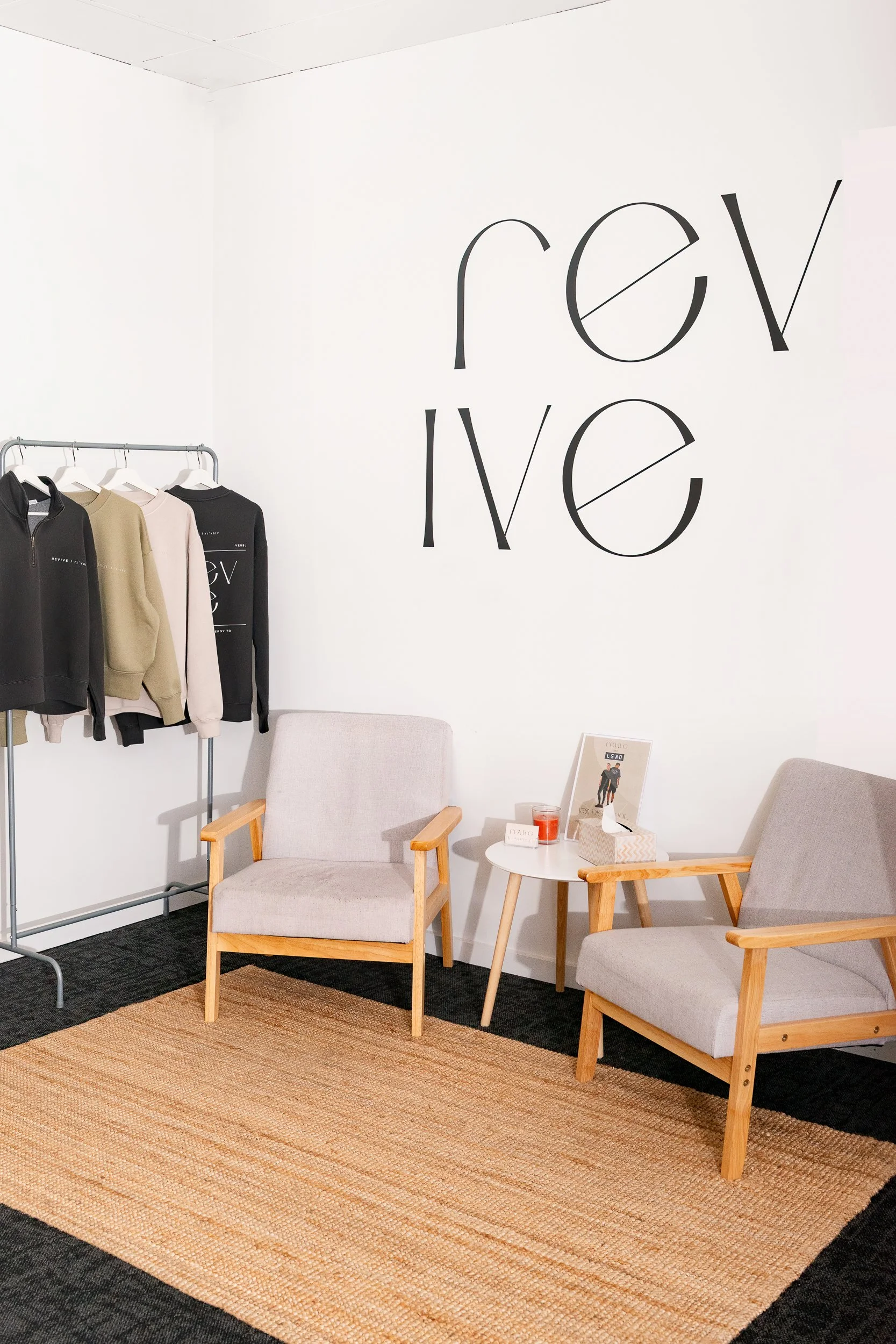 A cozy waiting area with two beige armchairs with wooden frames, a small round white table between them holding a framed photo, a tissue box, a candle, and a drink. To the left, a black clothing rack displays three hoodies in black, beige, and light pink with minimalist designs. The wall behind reads 'revis' in large black letters with a diagonal line crossing the 'e' and the 'v' in a modern font. The floor has a woven rug over a dark carpet.