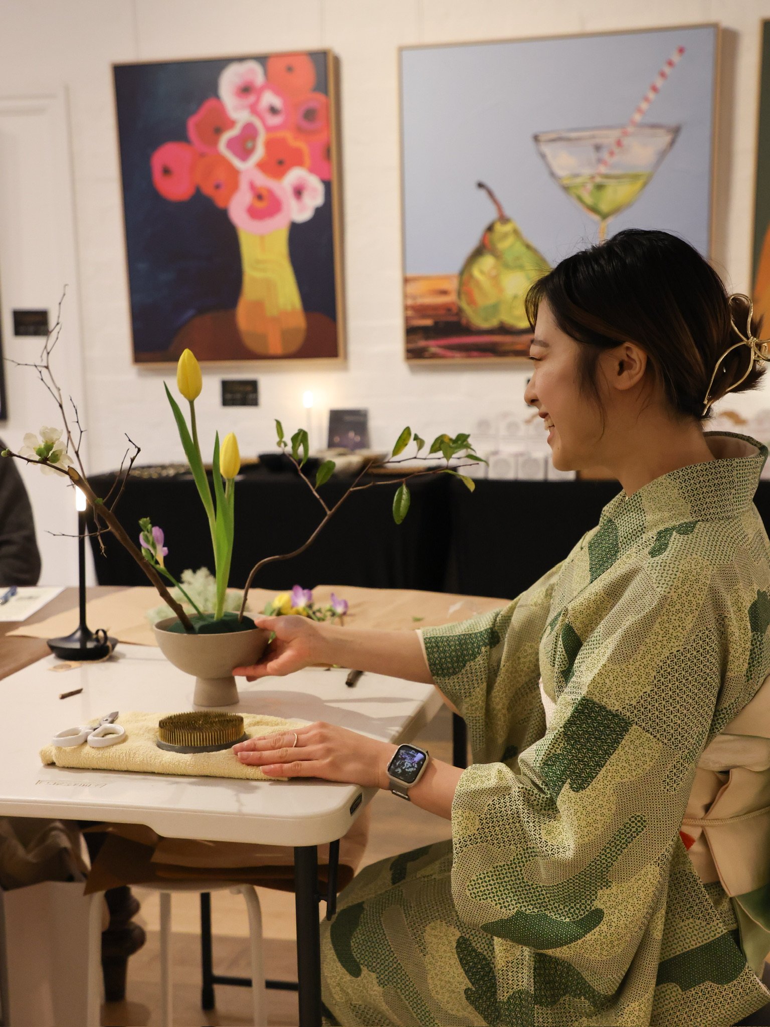 Person in green patterned kimono arranging flowers in a vase at a table, two paintings on wall, wearing a wristwatch.