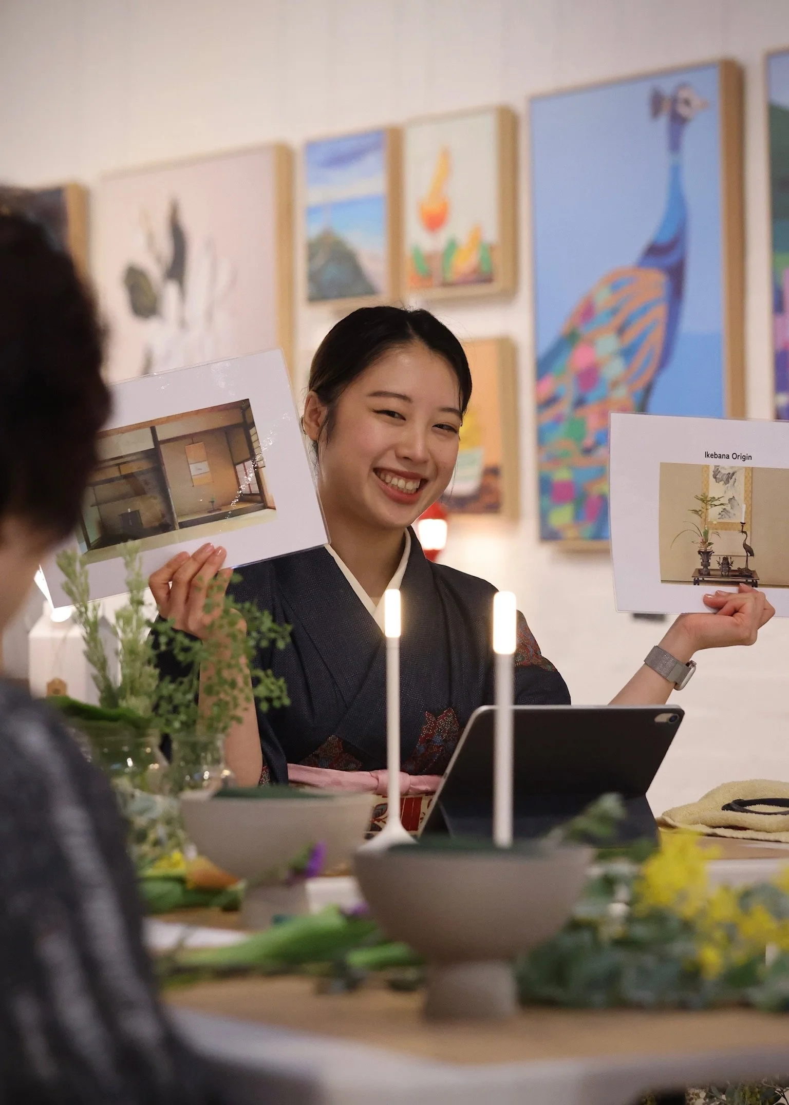 A woman in traditional attire is smiling and holding up two printed images during a presentation in a room with colorful artwork on the walls. There are flowers and lit candles on the table in front of her.