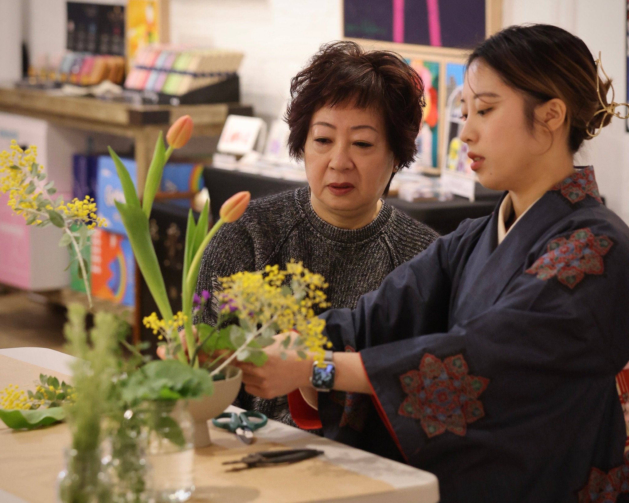 Two women arranging flowers at a table, one in traditional attire and the other in a sweater, with colorful flowers and tools visible.