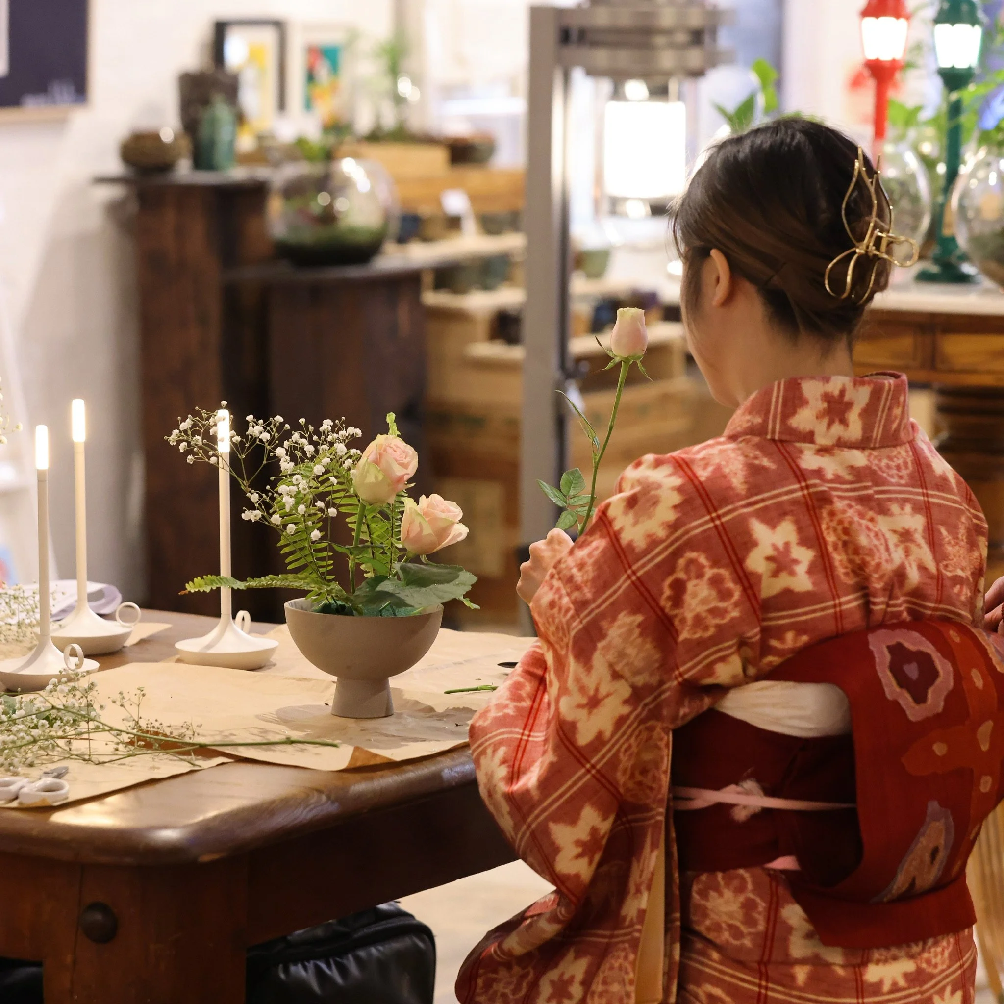 Person in red kimono arranging flowers at a wooden table, with candles and floral decorations in the background.