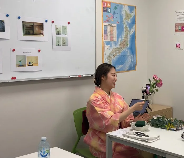 A woman in a traditional kimono demonstrates flower arranging at a table. A whiteboard and map are visible in the background.
