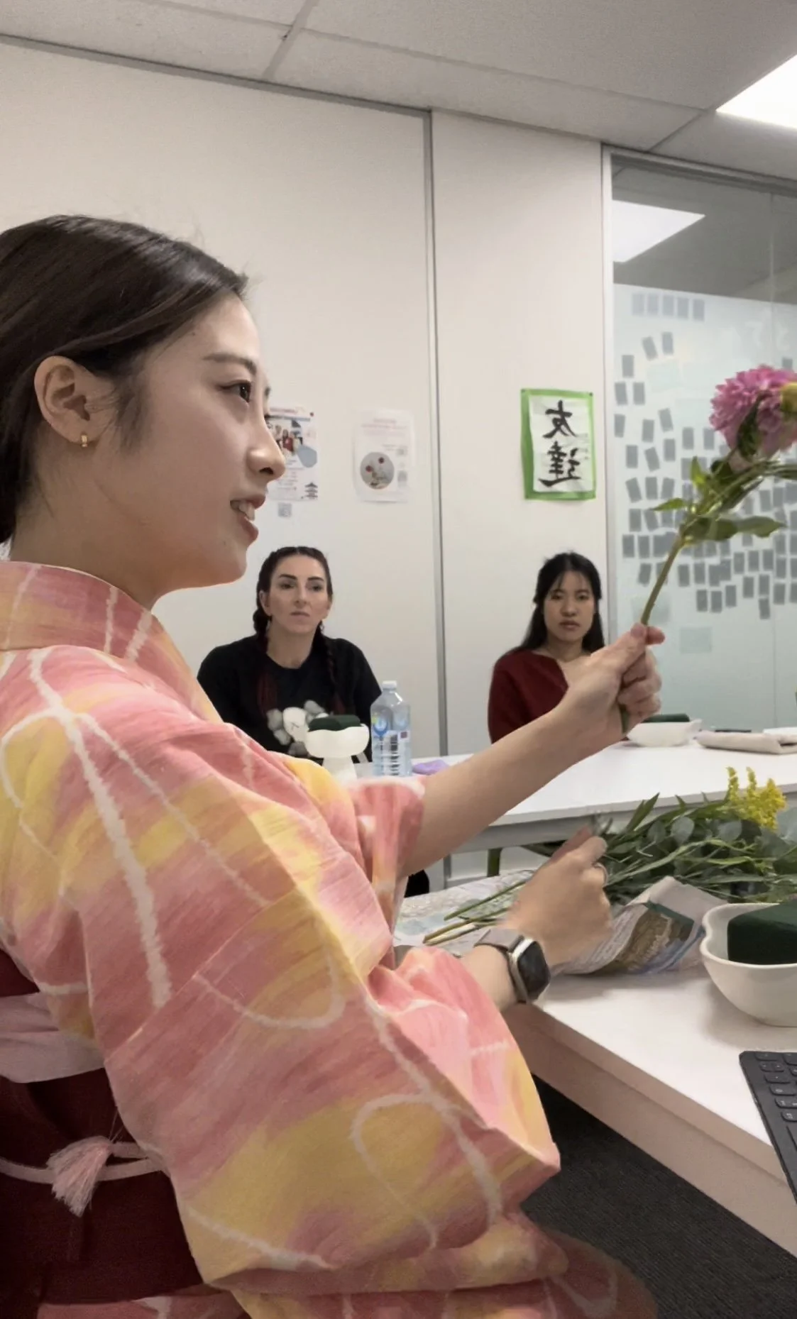 Woman in traditional Japanese attire holding flowers, seated at a table with other people.