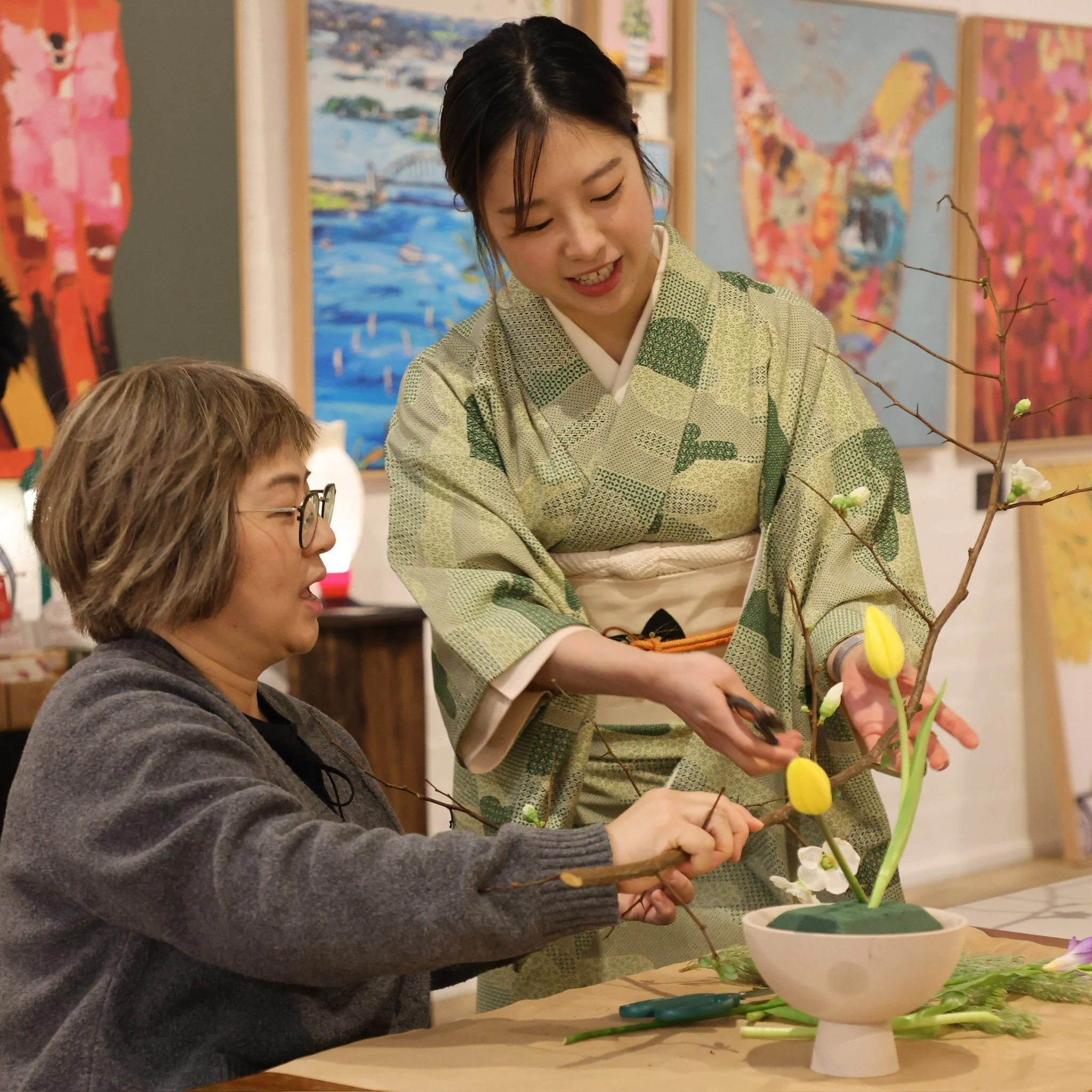 Two women arranging flowers in an ikebana workshop. One wears a kimono, assisting the other in arranging yellow tulips and branches in a bowl. Various colorful paintings are visible in the background.