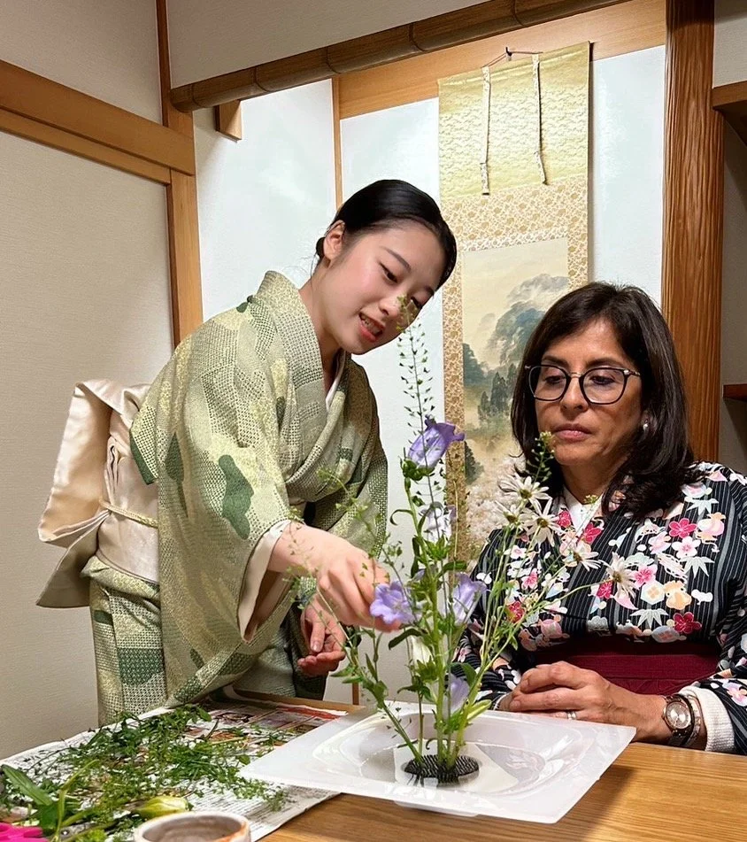 Two women in traditional kimonos engaged in flower arranging, known as ikebana, in a Japanese-style room with a hanging scroll.