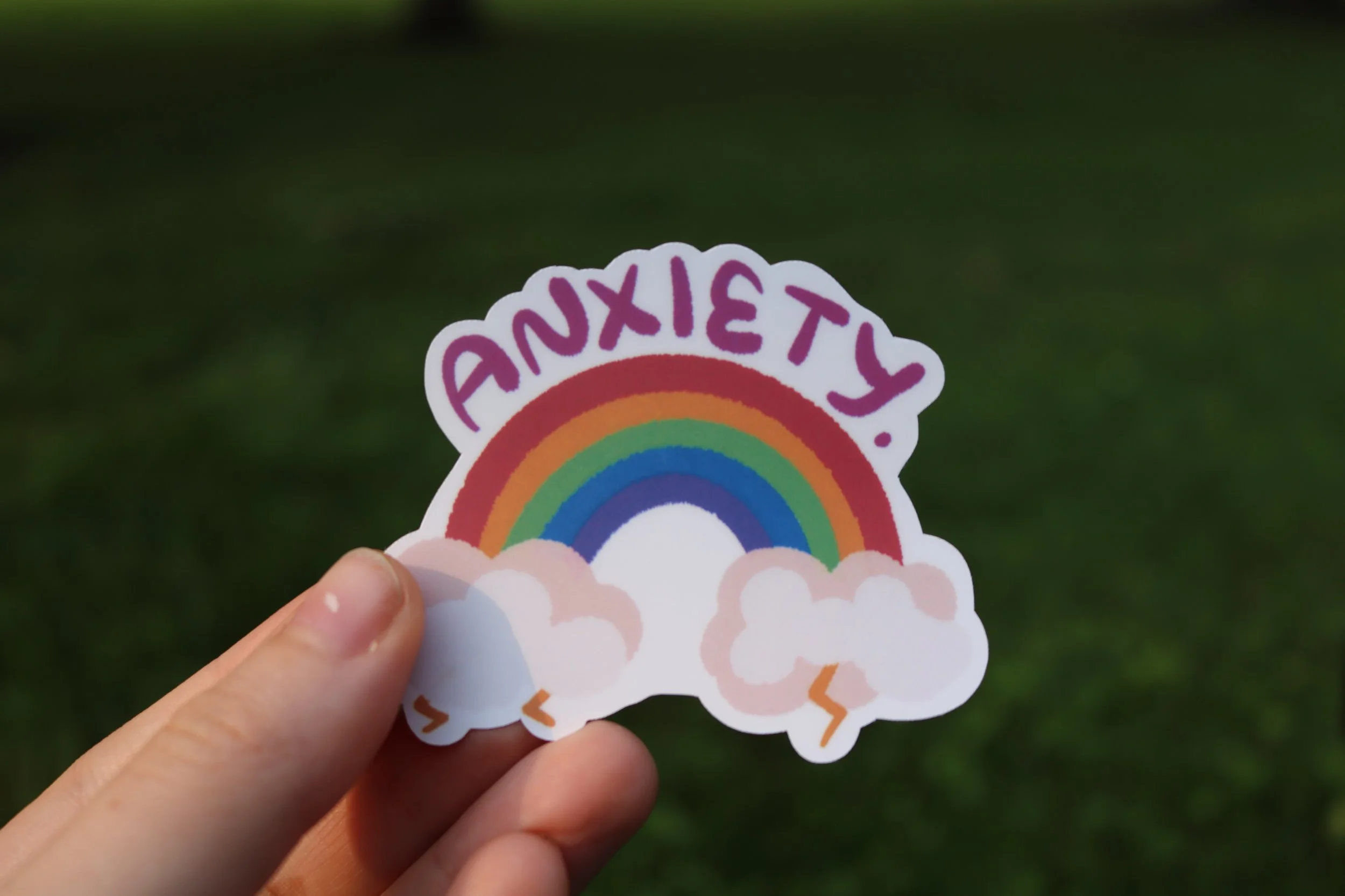 Hand holding a sticker of a rainbow with clouds and the words 'STAY SXUND' written above it.