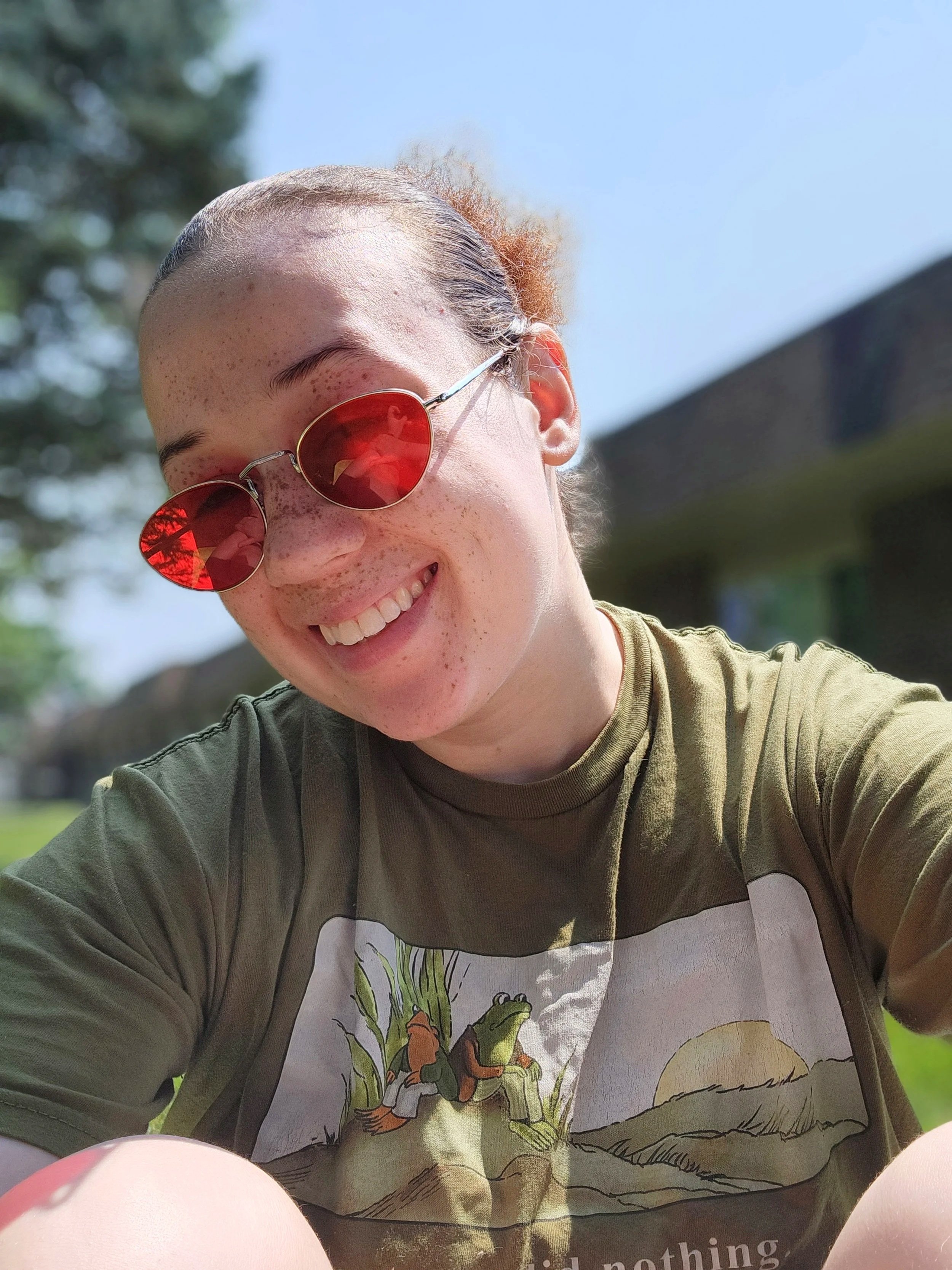 A person with a shaved head, freckles, and sunglasses smiling outdoors in bright sunlight. They are wearing a T-shirt with a graphic of a cartoon character and a green jacket, sitting on the ground with a blurred background of trees and a building.