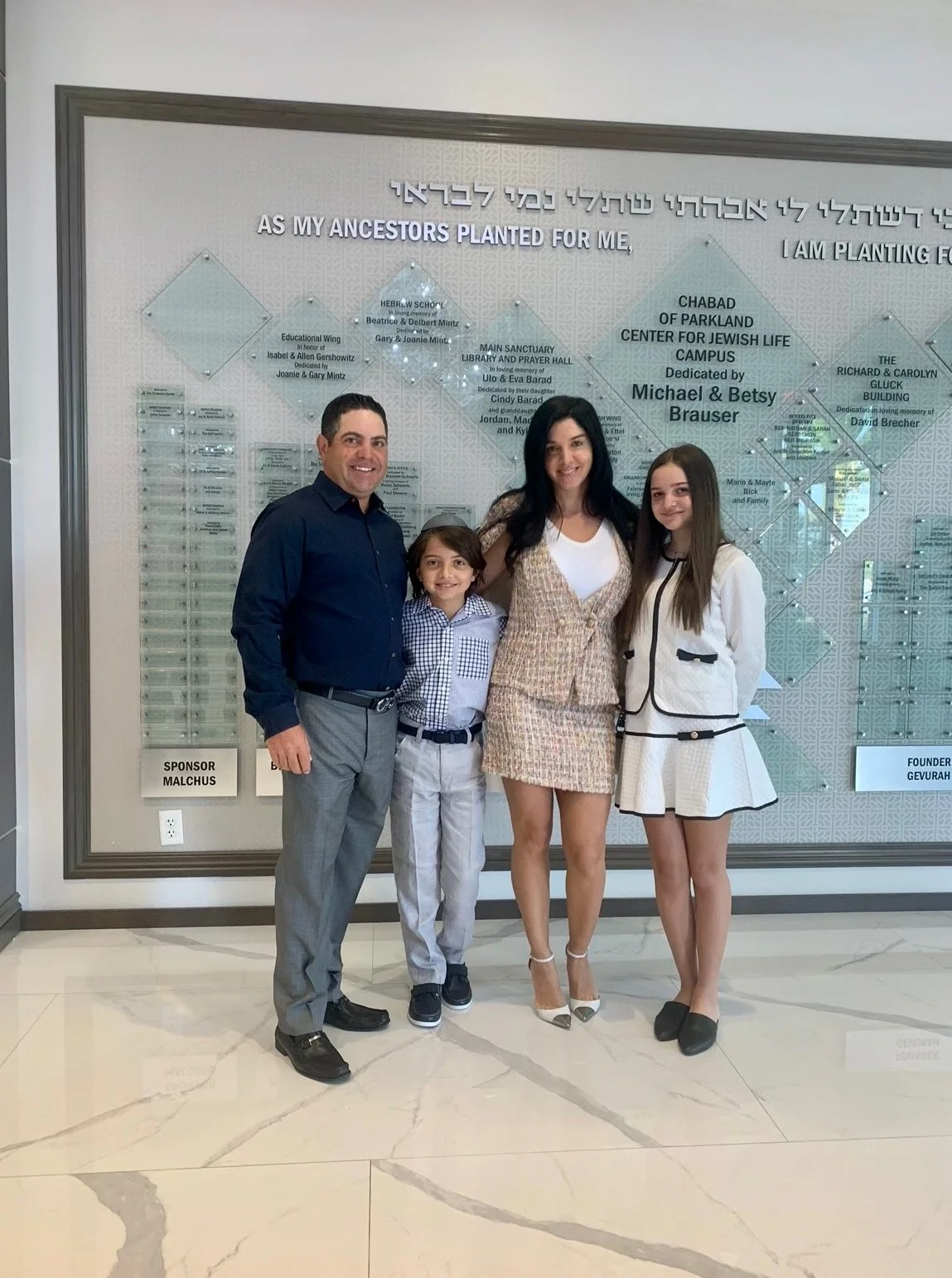 Daniel Kandler. Family of four standing inside a building in front of a wall with a large memorial plaque with Hebrew and English inscriptions. The family includes a man, woman, and two children, all smiling and dressed in semi-formal attire.