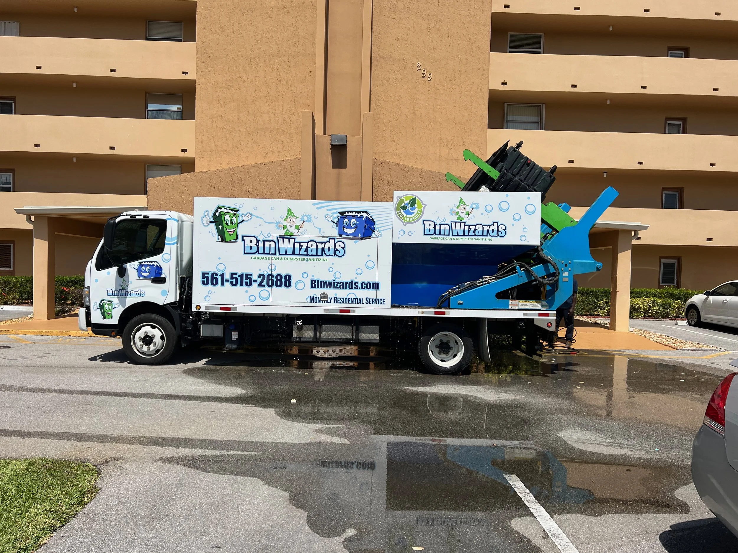 Daniel Kandler. A garbage truck with branding for Bin Wizards that sanitizes garbage cans and dumpsters, parked on a wet street in front of a residential building.