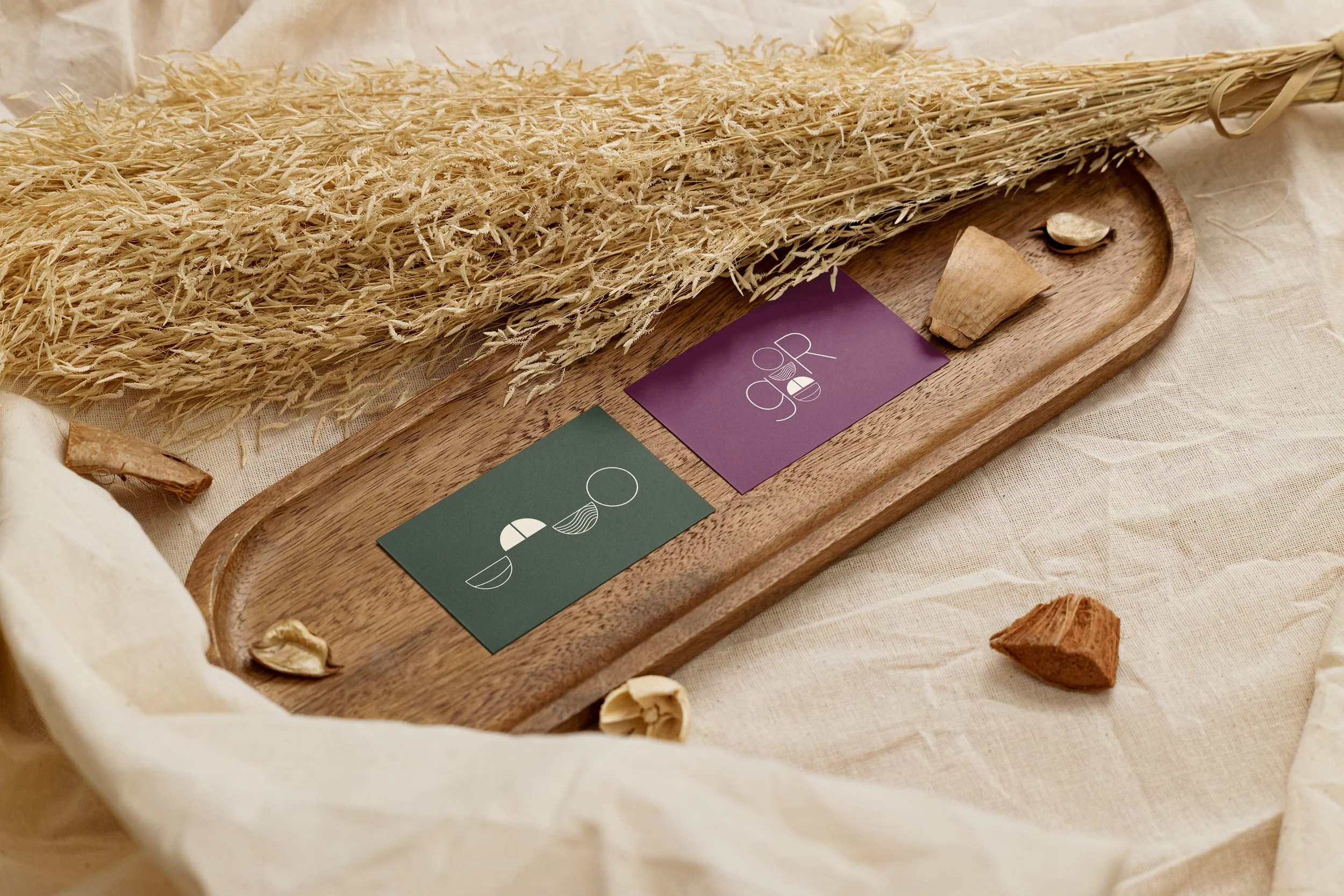 Zwei Visitenkarten mit minimalistischen Designs, ein Stück Holz und kleine Steine auf einem Holztablett, umgeben von getrocknetem Stroh und auf cremefarbenem Stoff