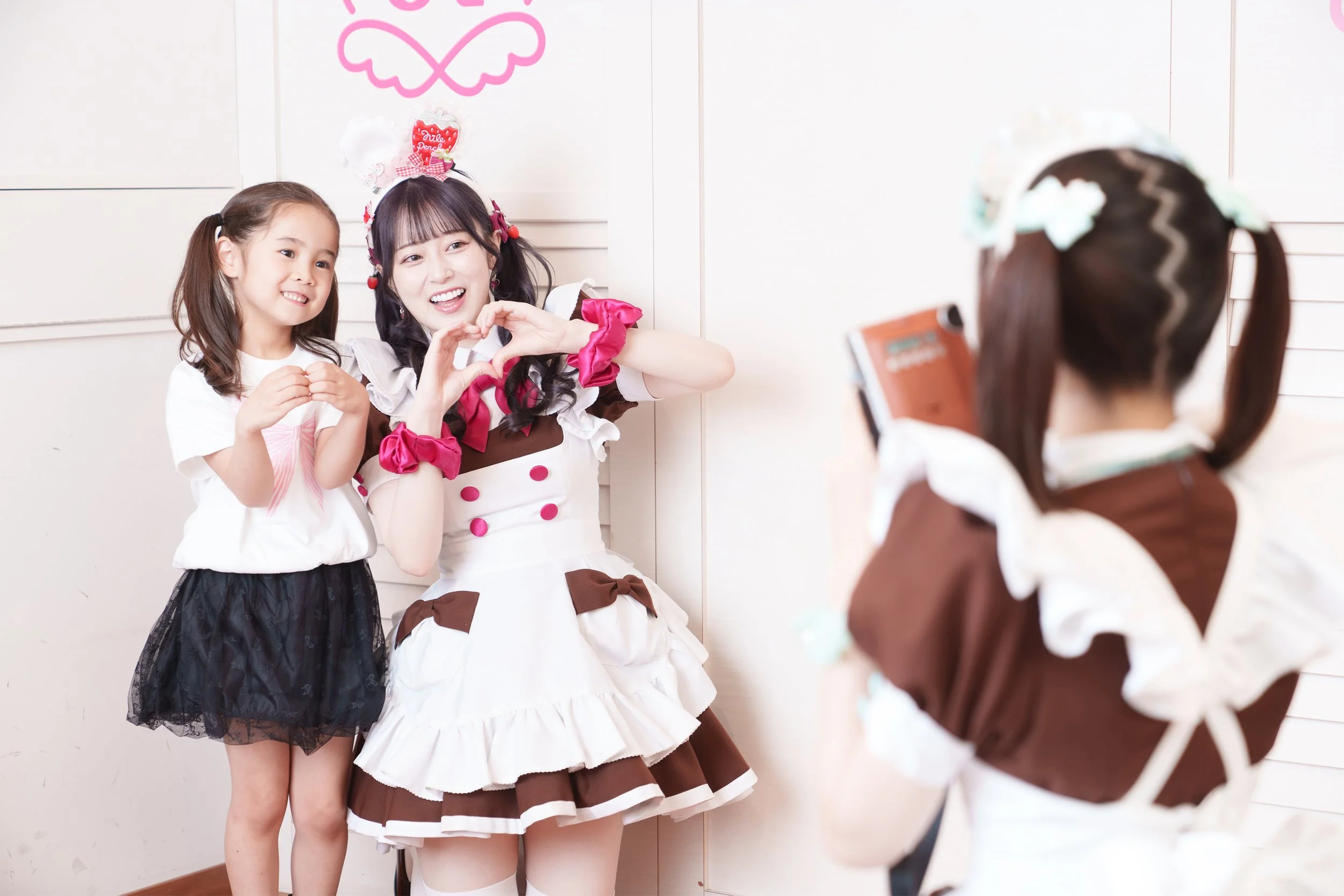 A woman and a young girl taking a photo of another girl dressed in a brown and white maid costume, making heart shapes with their hands at a party.