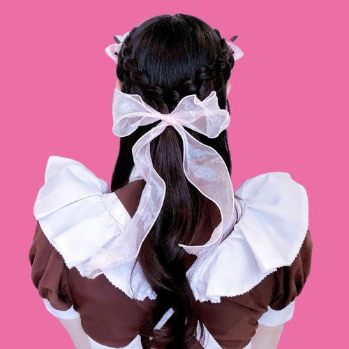 Back view of a girl with long dark hair styled in braids, wearing a white and brown dress with puffed sleeves, and a large white bow tied in her hair against a pink background.