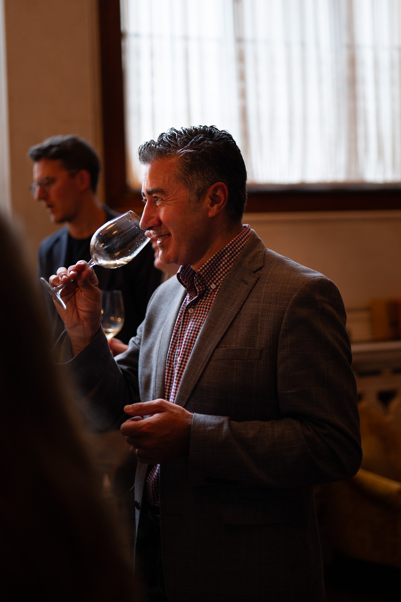 Man smiling and smelling a glass of white wine during a social event, with a blurred man in the background.